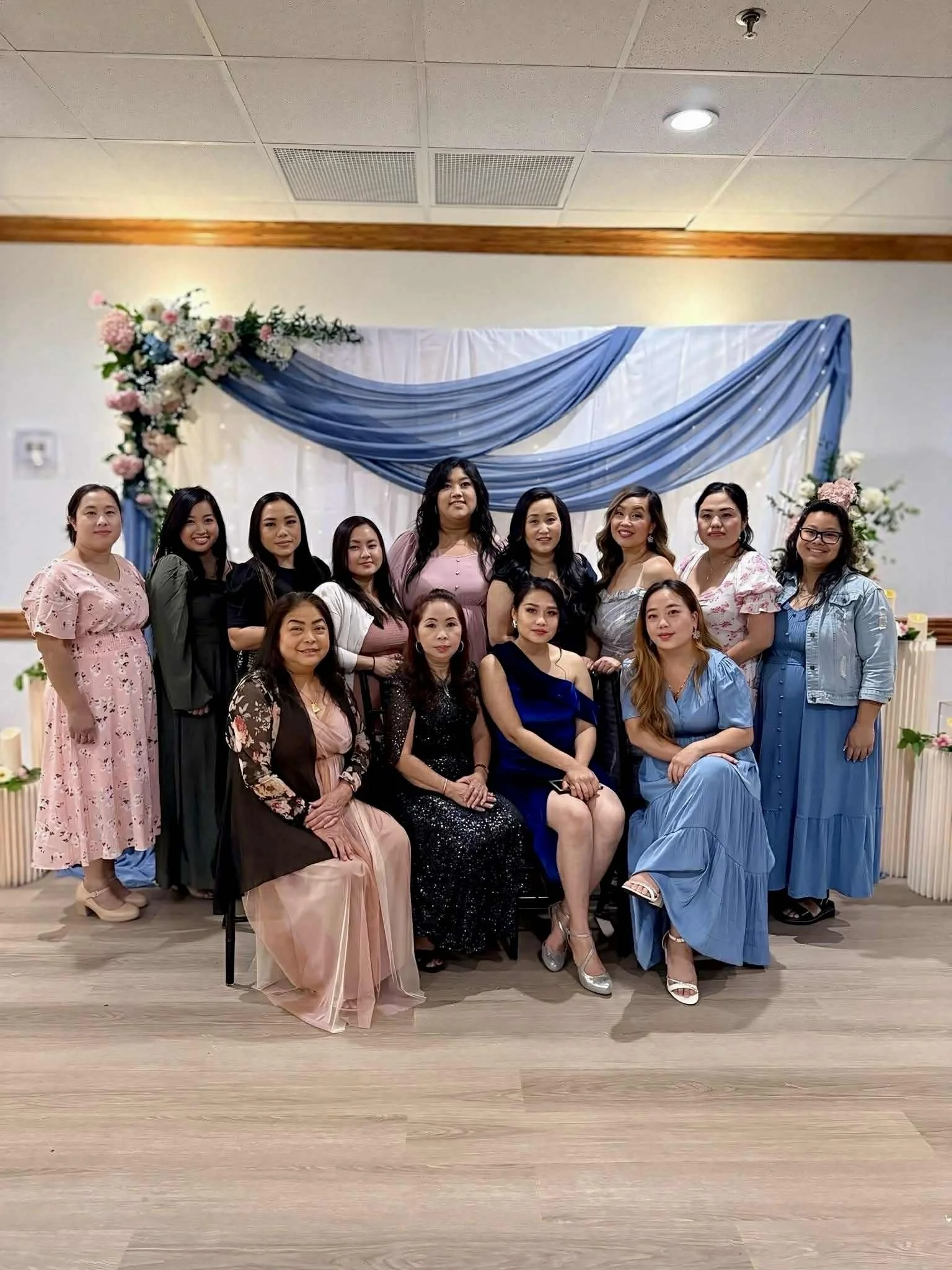 A group of women in formal dresses posing for a photo in front of a decorated backdrop with blue drapes and floral arrangements.