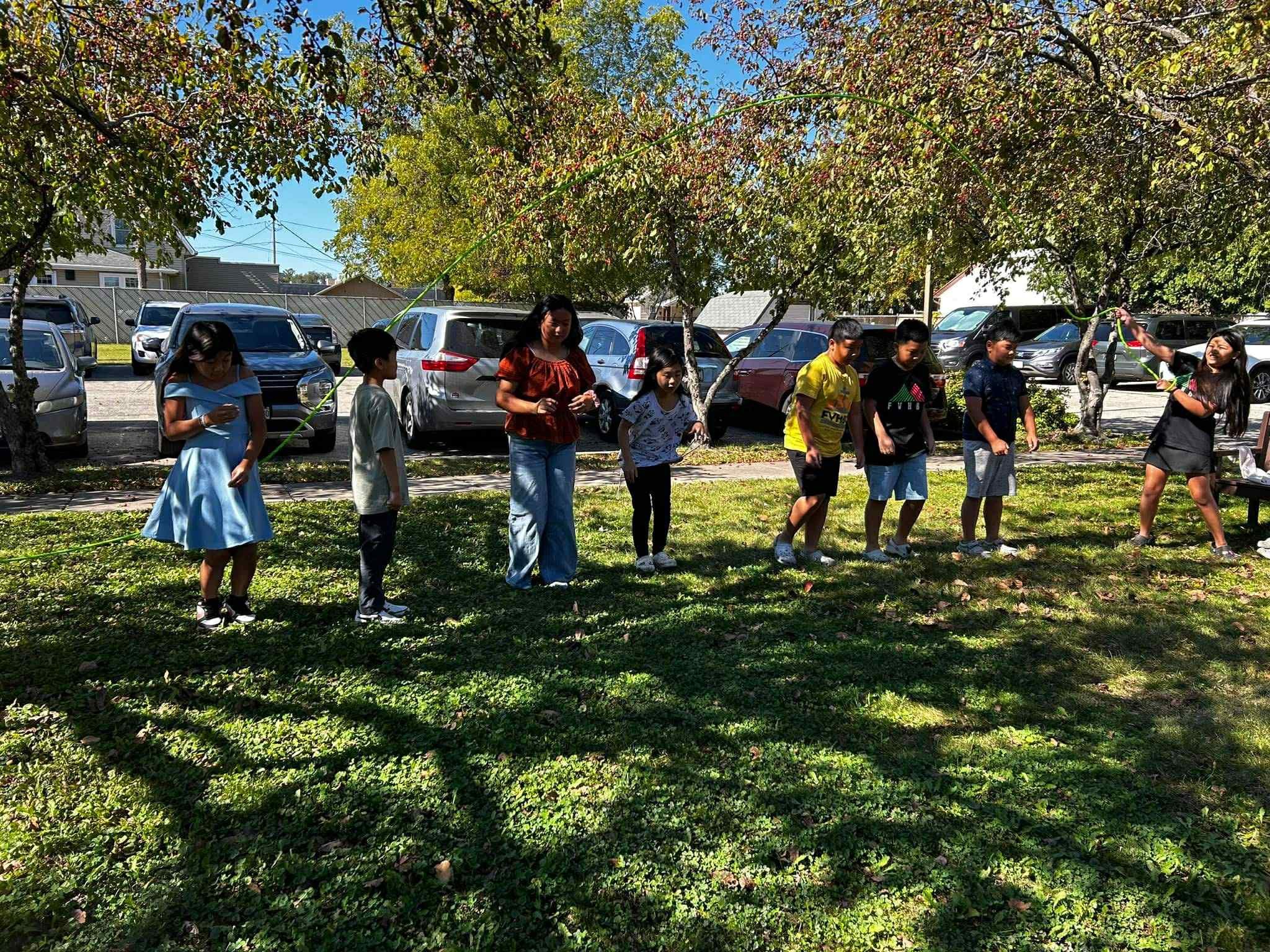Children and adults playing jump rope in a park with trees and parked cars in the background.