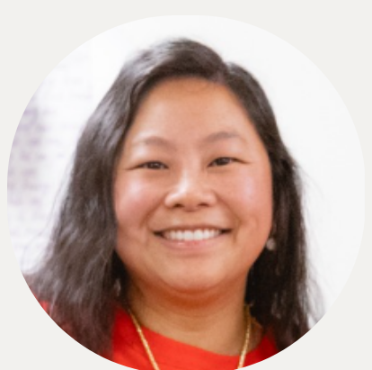 A woman with long, dark hair wearing a black blazer and green t-shirt, standing against a plain white wall.