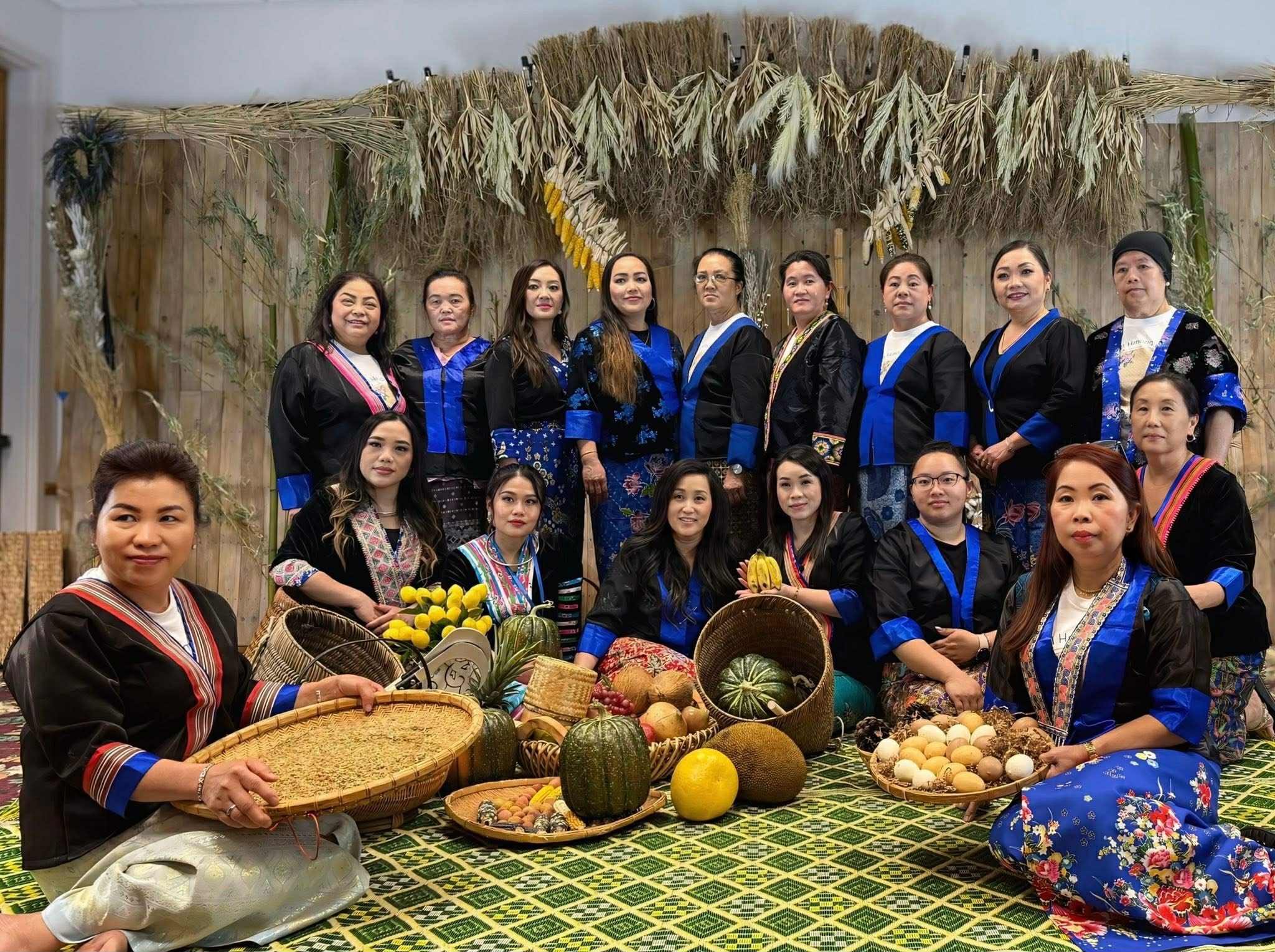 A group of women dressed in traditional clothing, sitting and standing around a display of seasonal fruits and vegetables, with a decorated straw and plant backdrop.