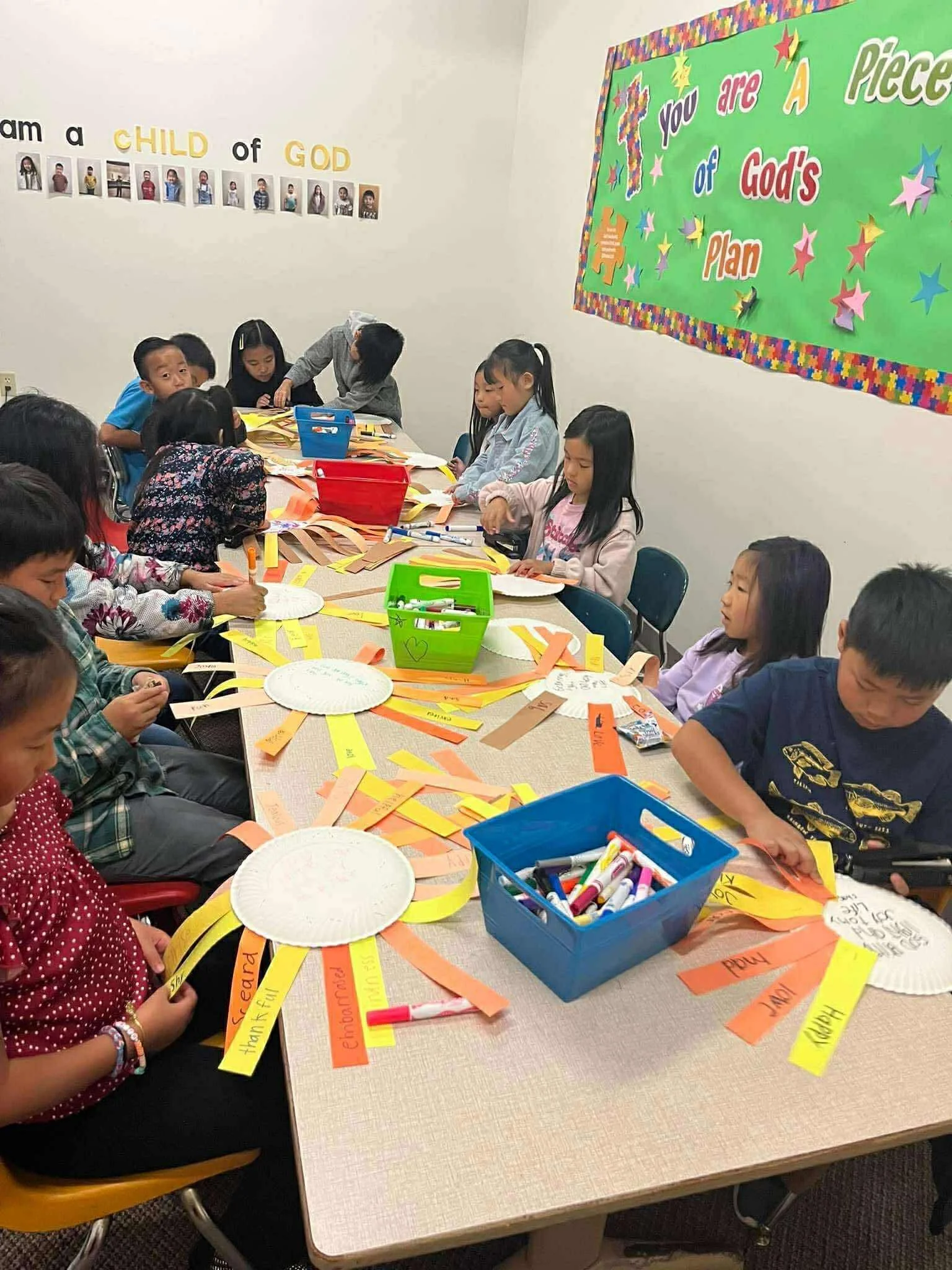 Children sitting around a table engaging in arts and crafts activities, using paper, markers, and scissors in a classroom decorated with educational posters and colorful wall art.