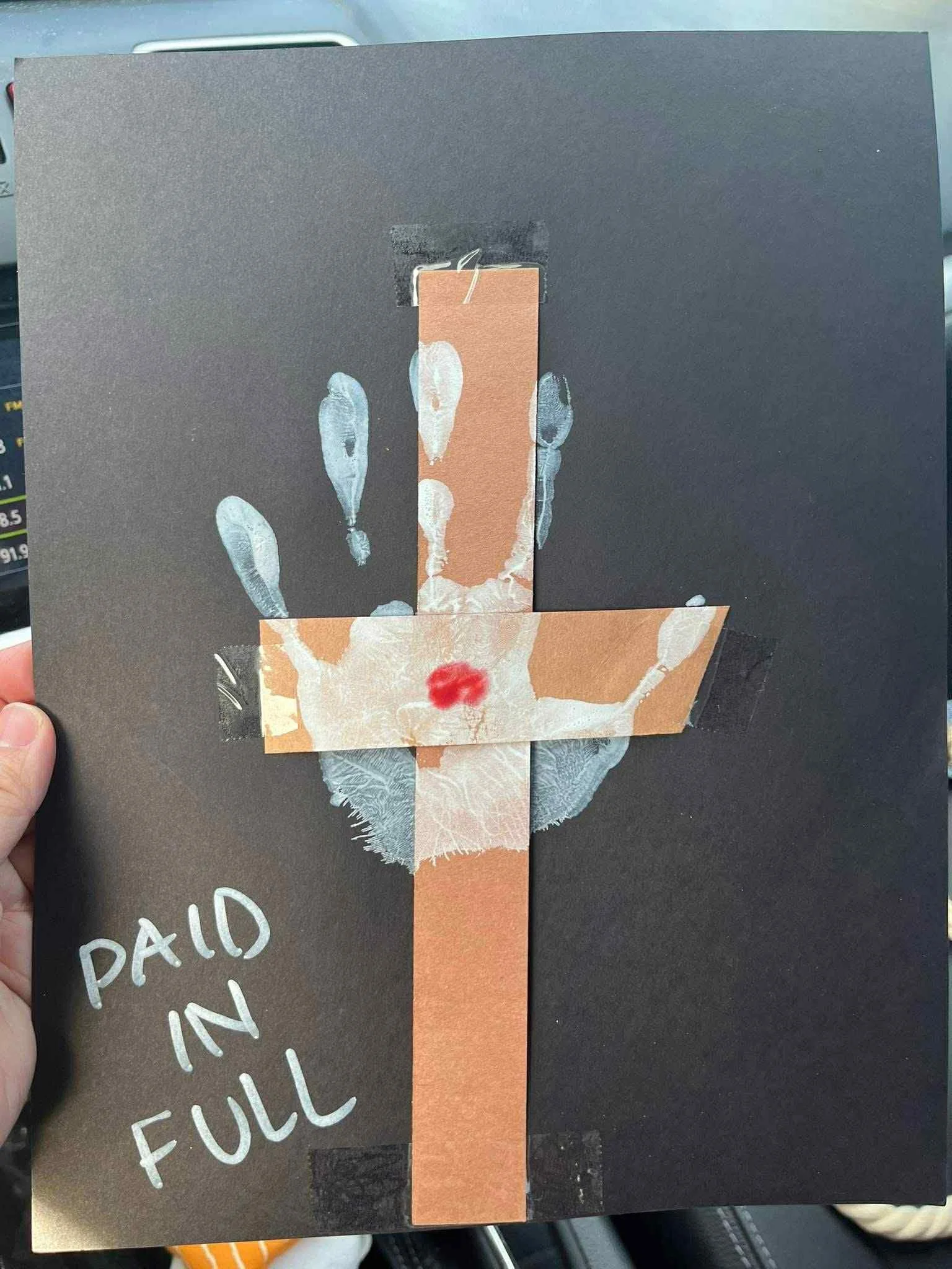 A black poster with a collage of various paper cutouts forming a cross, featuring a white handprint with a red lipstick mark on the palm, the words "PAID IN FULL" written in silver marker, and some tape at the edges.