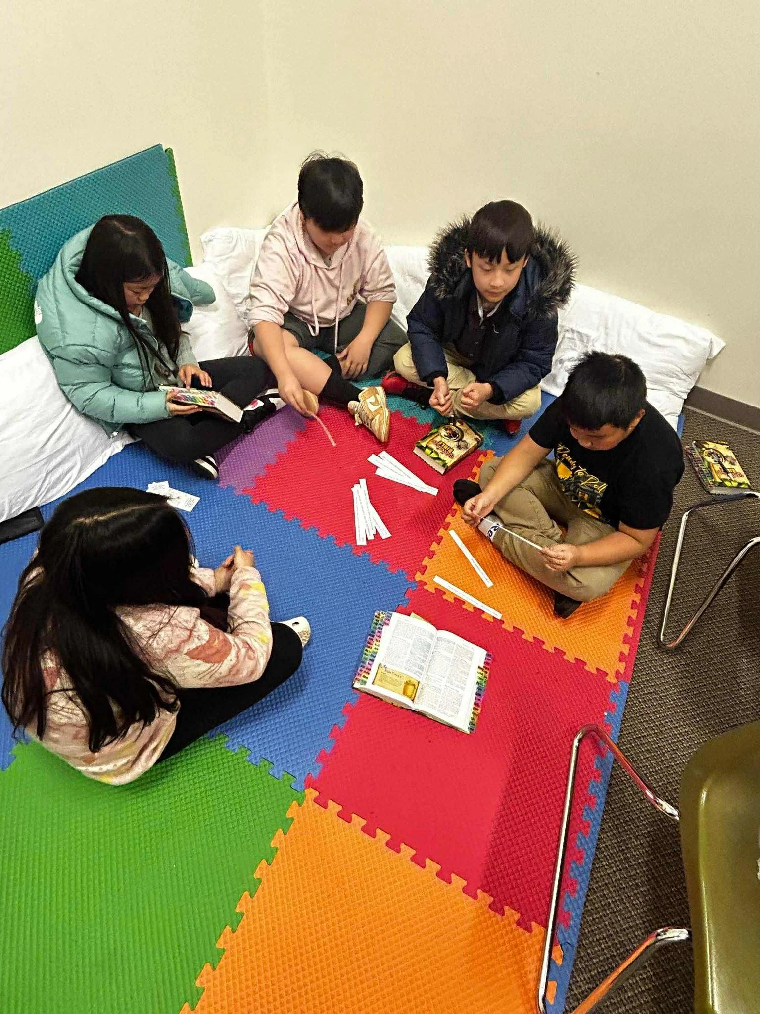 Five children sitting on a multicolored foam mat, reading books and playing with small sticks, in a cozy indoor setting with cushions and a small table nearby.
