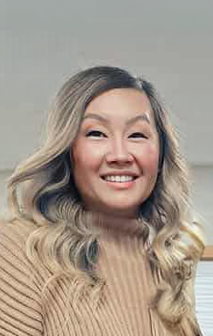 A woman with dark hair and wearing a floral dress, gray jacket, pearl necklace, and red necklace, standing indoors in front of a cross and music stand.