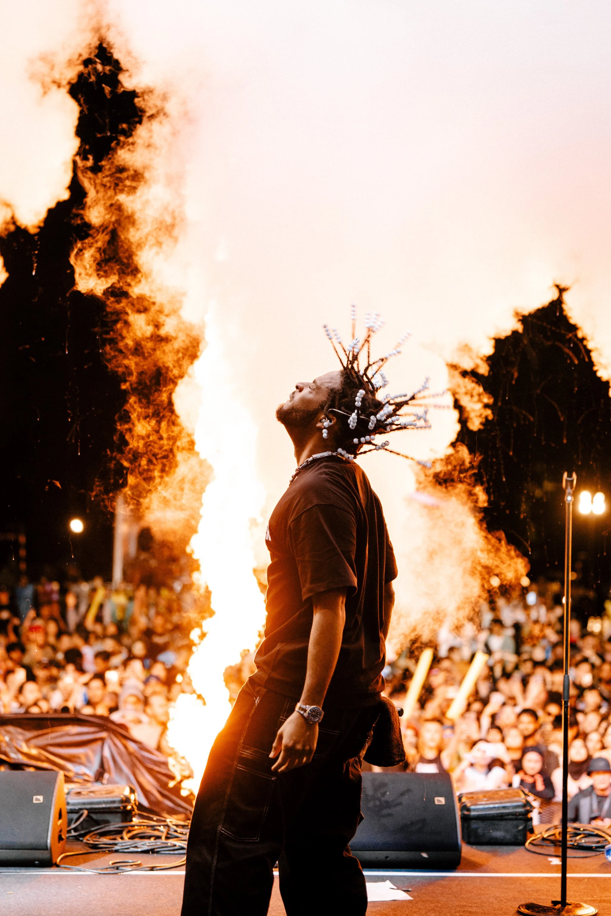 Performer with dreadlocks under fire during concert, with large audience in background.