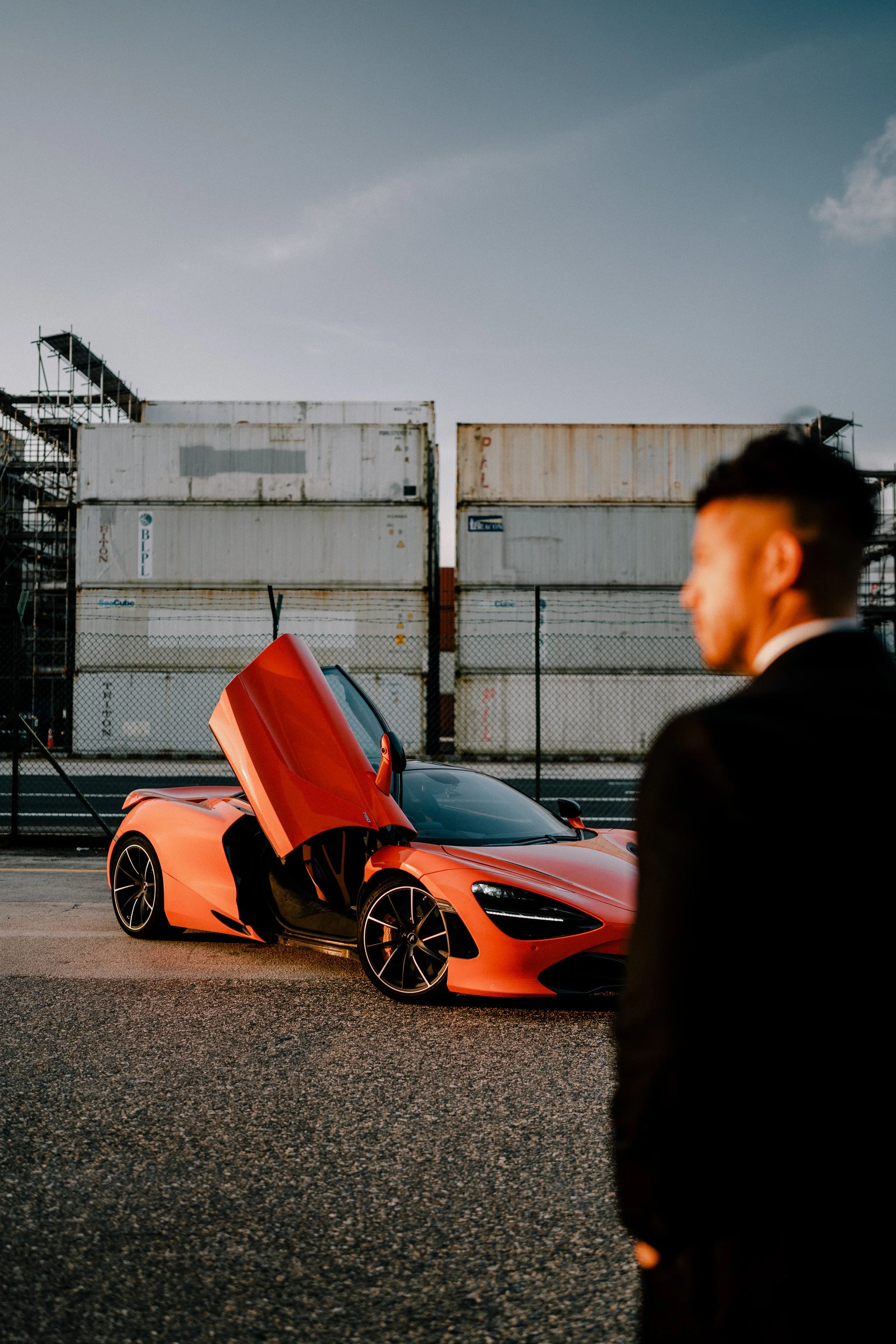 A bright orange sports car with its driver's side door open in an industrial area, a person in a black suit with a blurred face in the foreground, and shipping containers in the background.