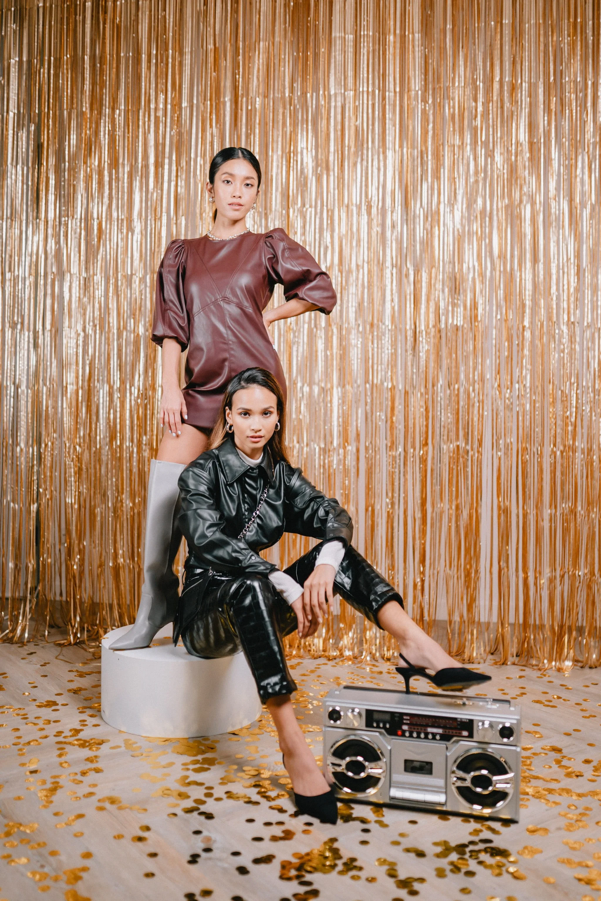 Two women in fashionable outfits pose in front of a gold foil curtain backdrop, with a boombox on the floor and scattered gold confetti.