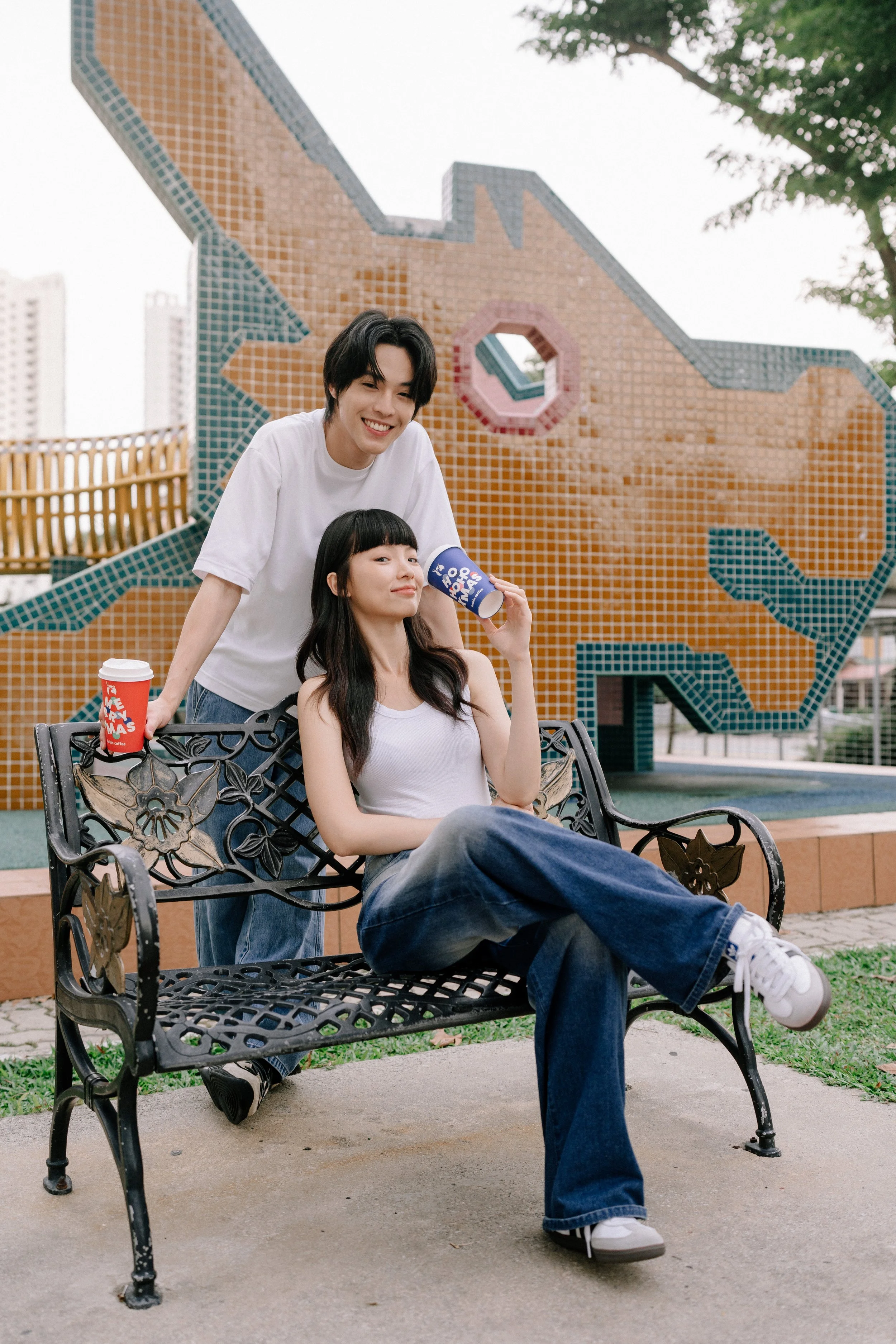 Two young women with dark hair, one sitting on a park bench and the other standing behind her, holding drinks, in front of a colorful mosaic wall with a geometric design, outdoors in a park.