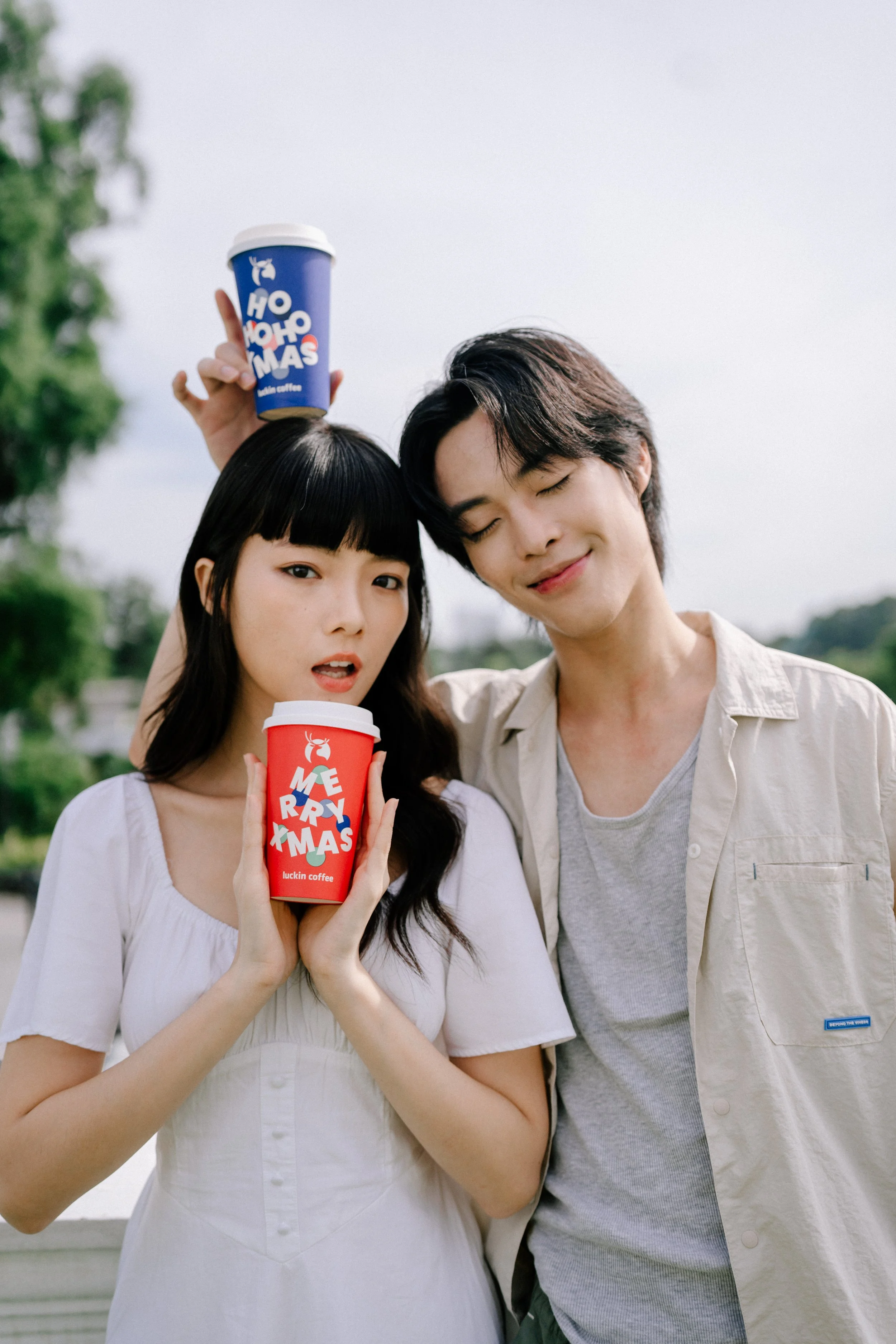 A young woman and a young man outdoors holding holiday-themed coffee cups, with the woman’s cup reading 'MERRY XMAS' and the man’s cup reading 'HO HO HO MERRY XMAS'.