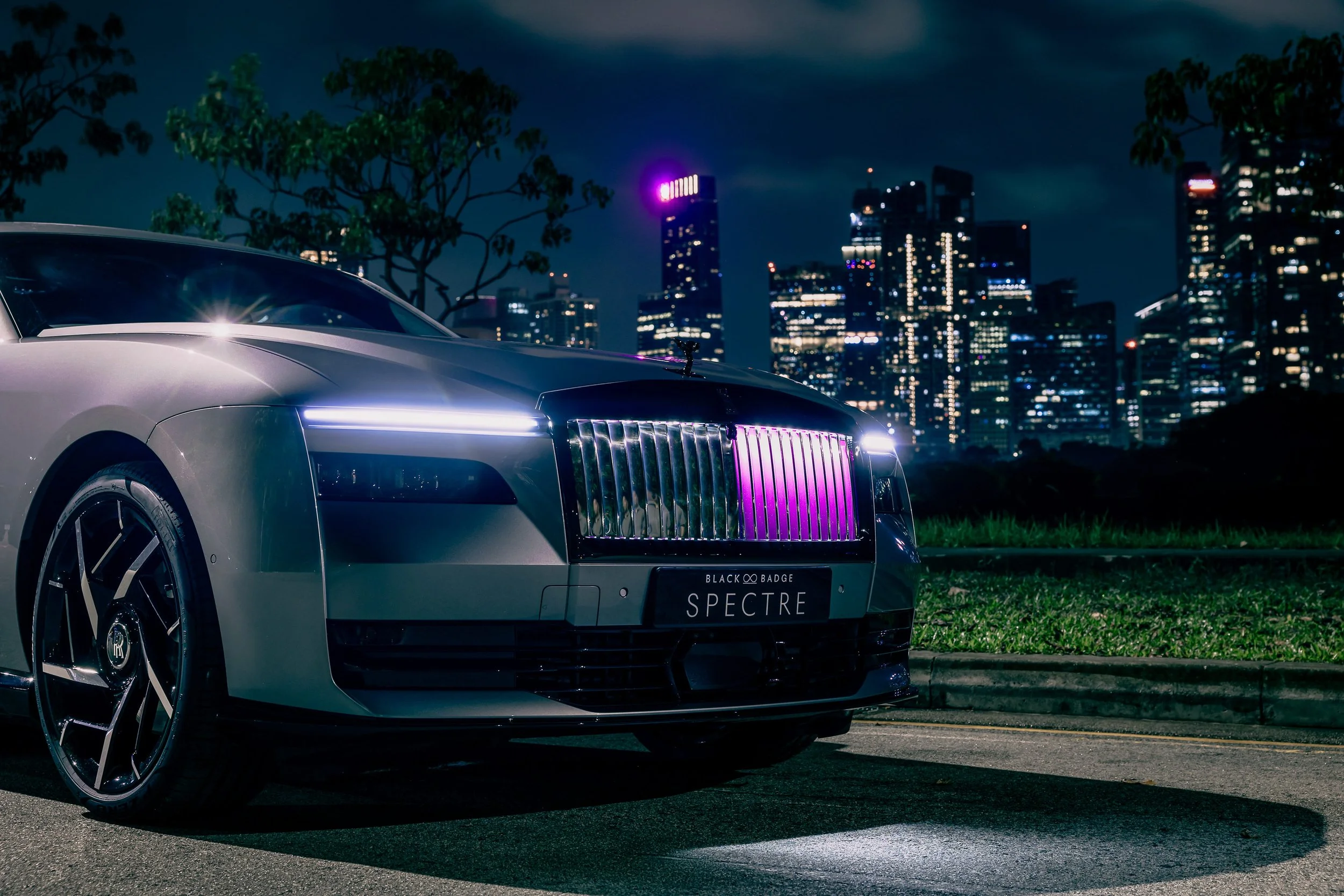A silver Rolls-Royce Spectre parked on a city street at night with a city skyline lit in the background, illuminated with purple and white lights.