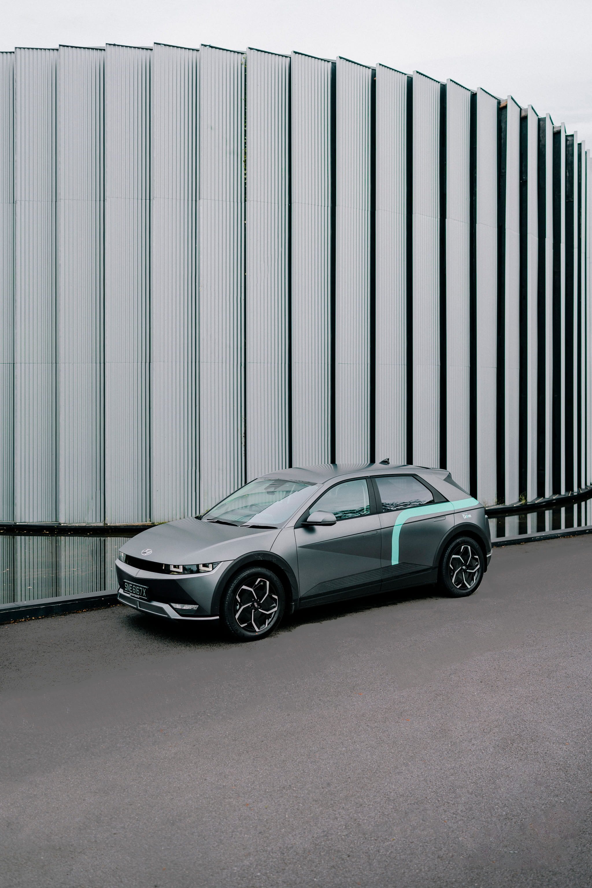 A modern, gray electric car parked beside a reflective water feature and a large metallic building with vertical panels.