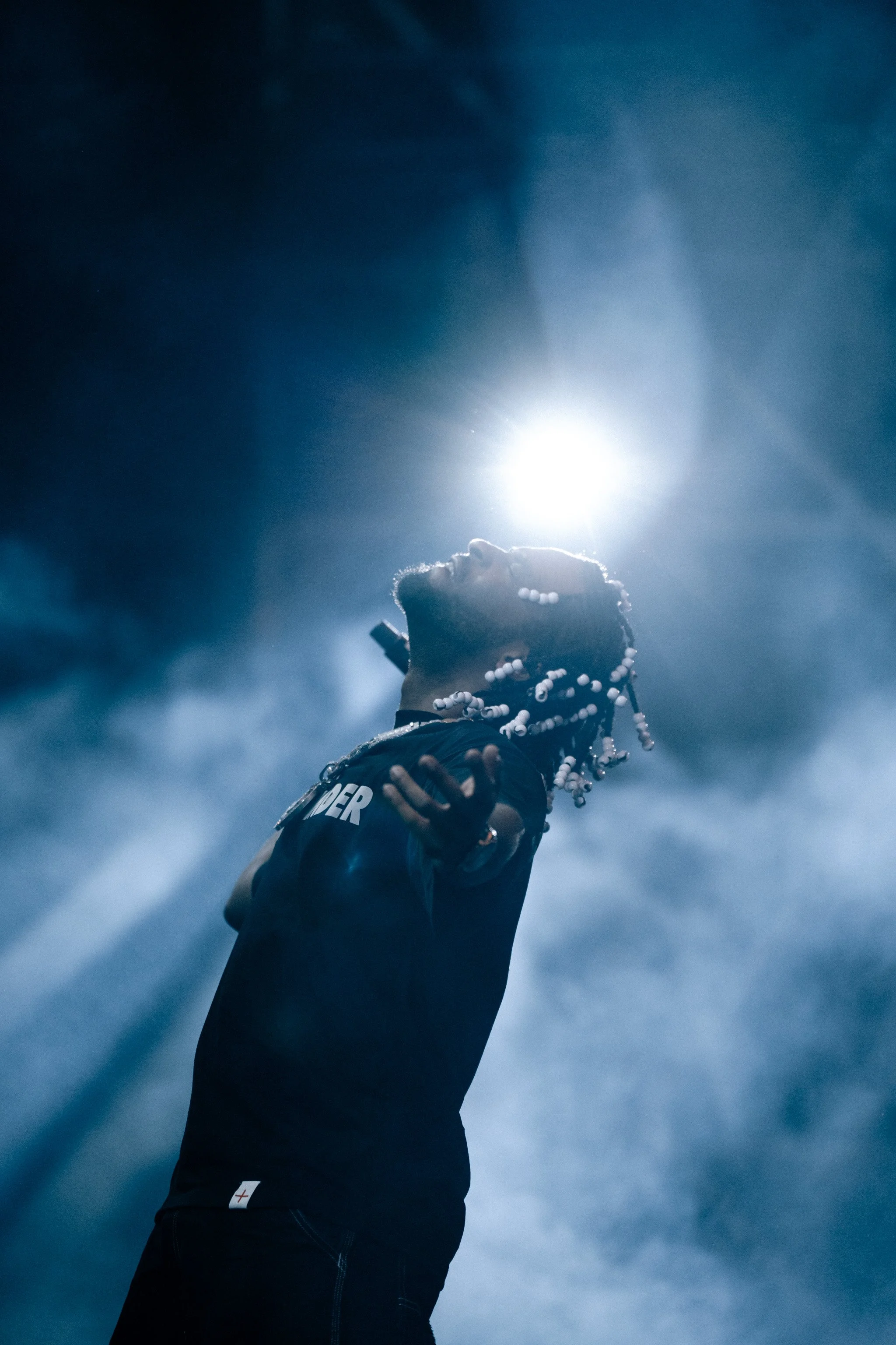 A man with beaded hair, wearing a black jacket, stands outdoors at night with a bright light behind him, surrounded by smoke or clouds.