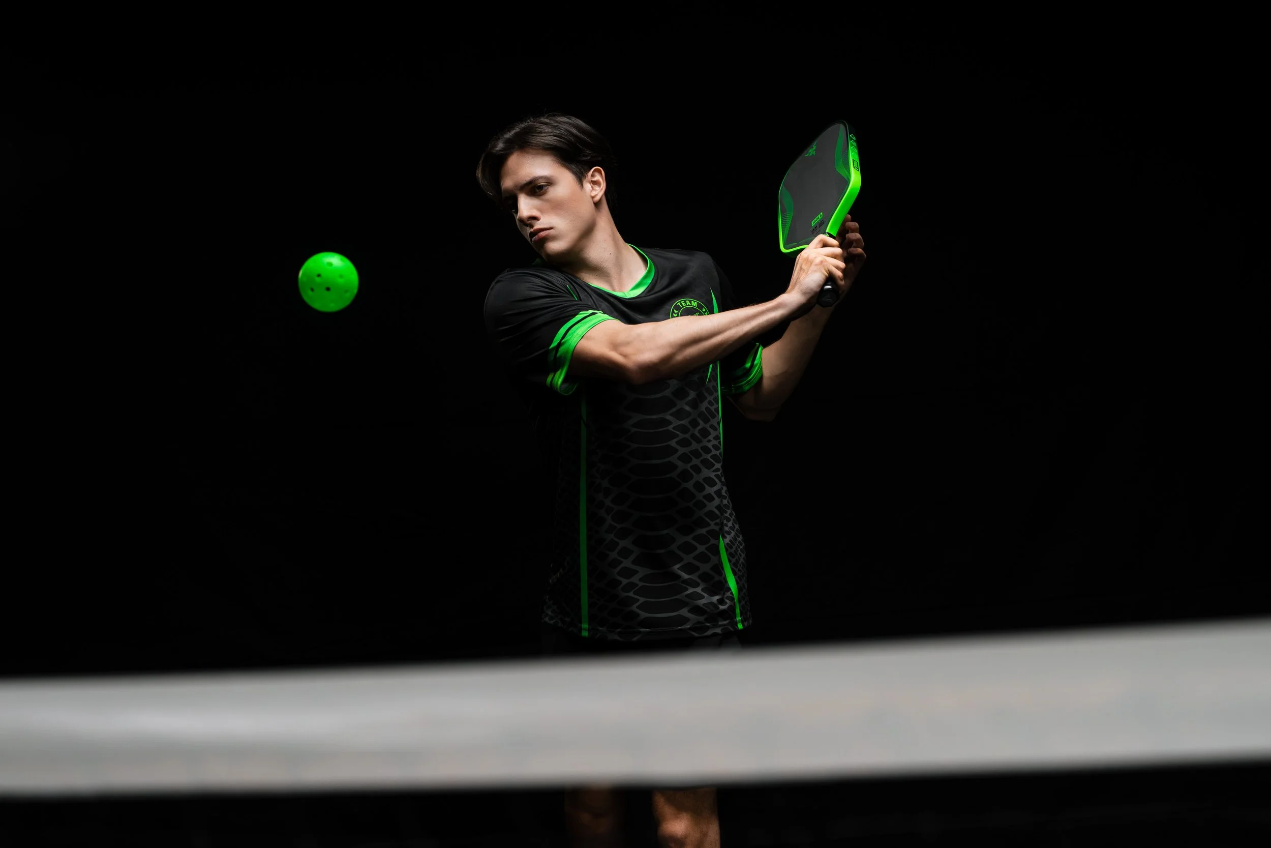 A young man playing pickleball, preparing to hit a green ball with a black and green paddle in a dark environment.