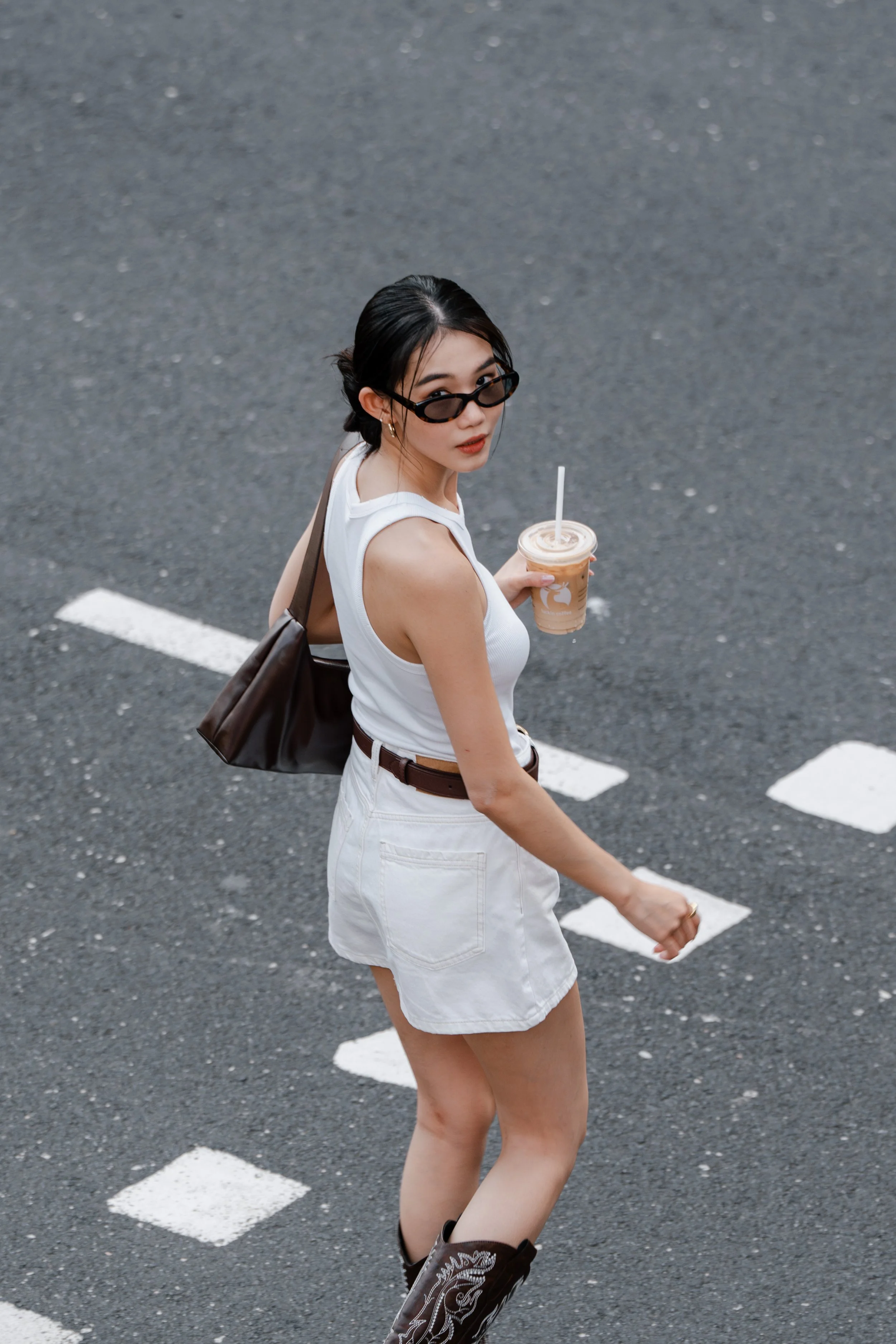 A woman in a white tank top and shorts, wearing sunglasses, walking on an asphalt street with white painted lines, holding a iced coffee drink.