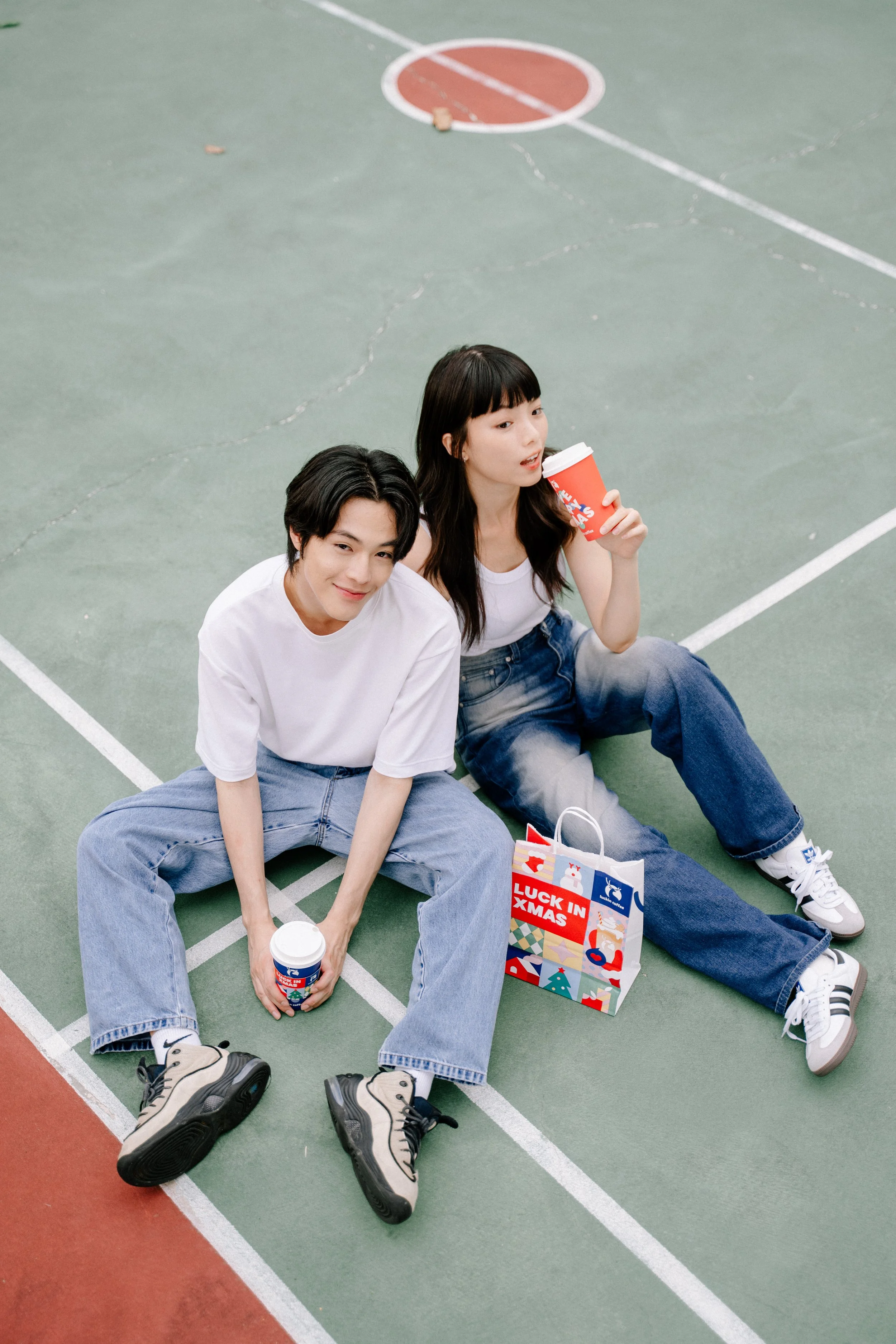 Two young people sitting on a basketball court, drinking from holiday-themed cups, with a Christmas gift bag nearby that says "Luck in Xmas."