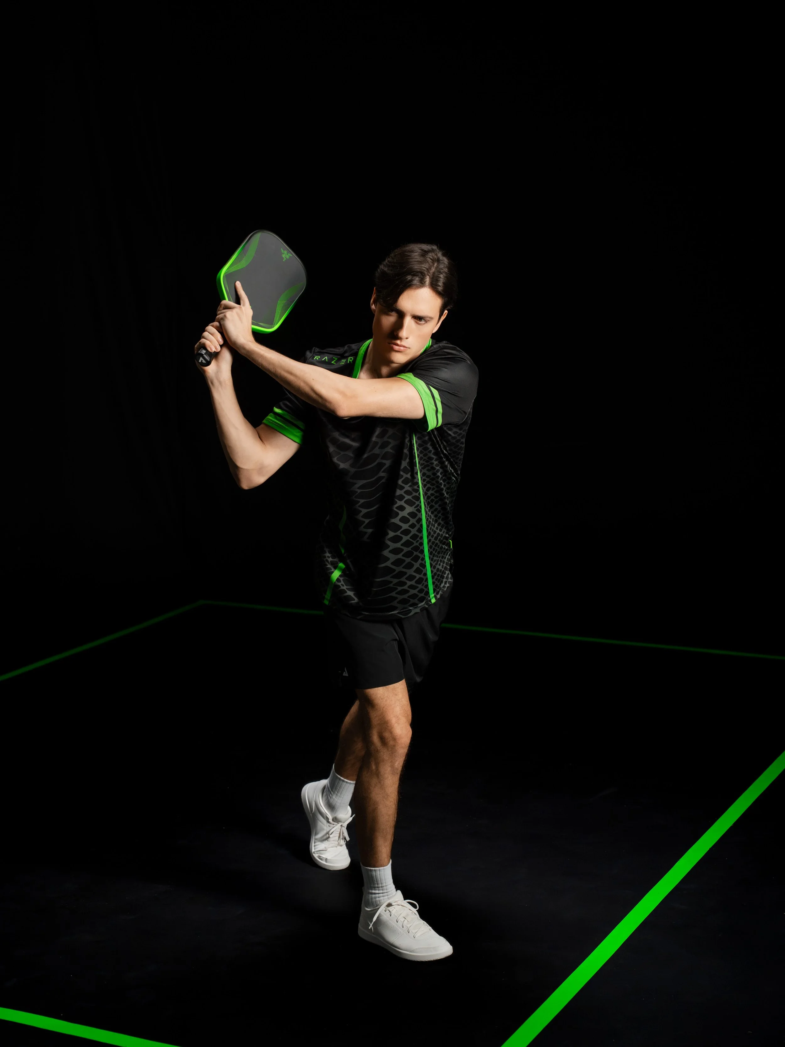 A young man in black and green athletic clothing prepares to hit a black and green Razer-branded paddle in a minimalistic black room with green accent lines on the floor.