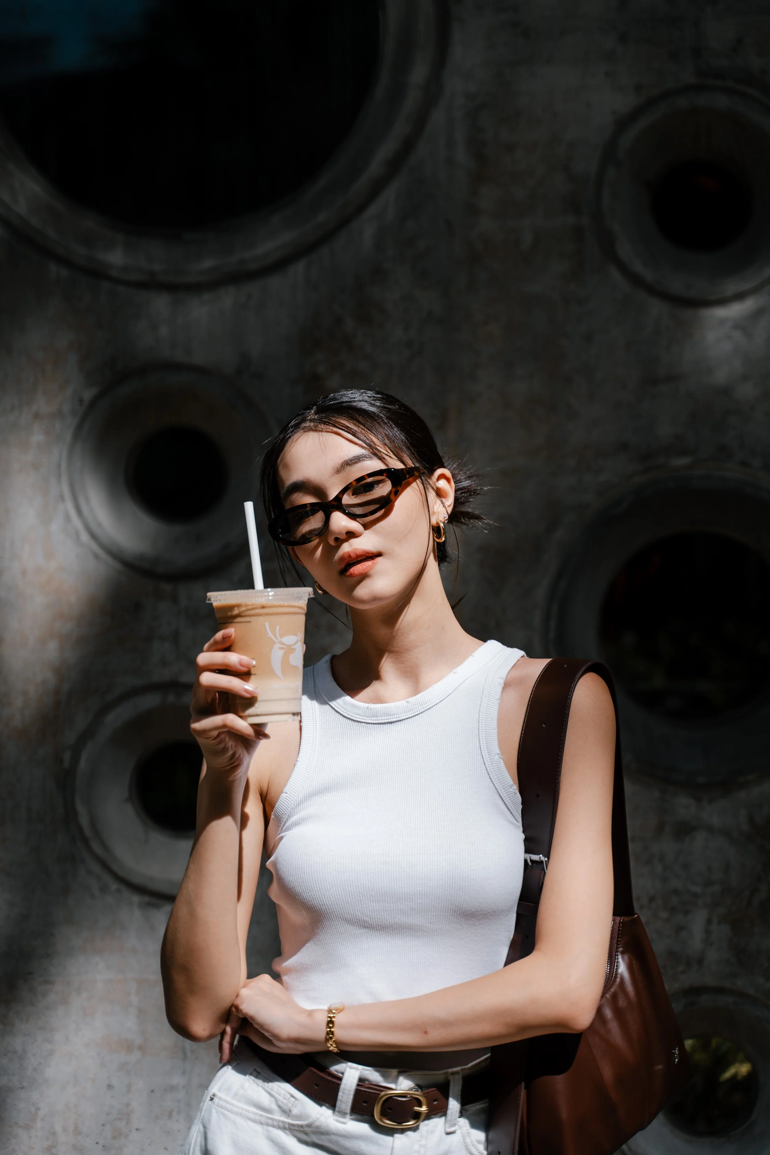 A young woman wearing sunglasses, a white sleeveless top, and a brown purse is holding an iced coffee with a straw. She is standing outdoors in front of a textured concrete wall with circular design elements.