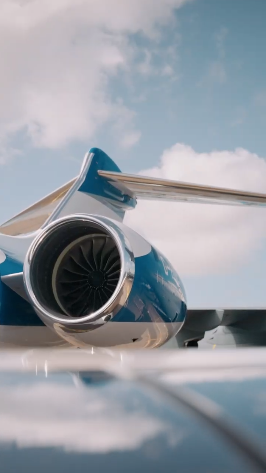 Close-up of the rear jet engine and tail section of a private jet aircraft with blue and white color scheme, against a partly cloudy sky.
