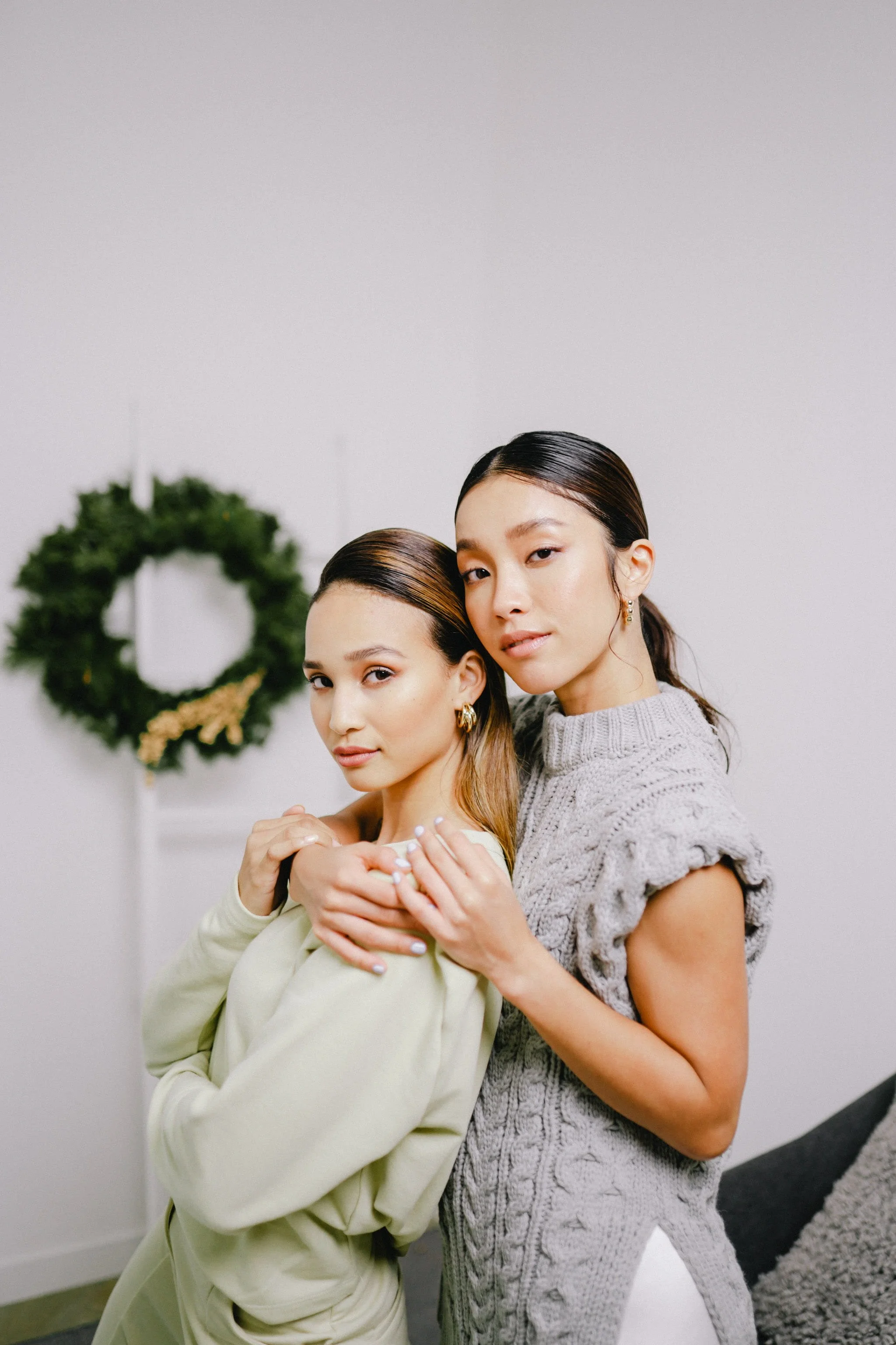 Two women standing close together, one with long dark hair and the other with shoulder-length hair, both looking at the camera with neutral expressions. The woman on the right has her arms around the other. A decorative holiday wreath hangs in the background.