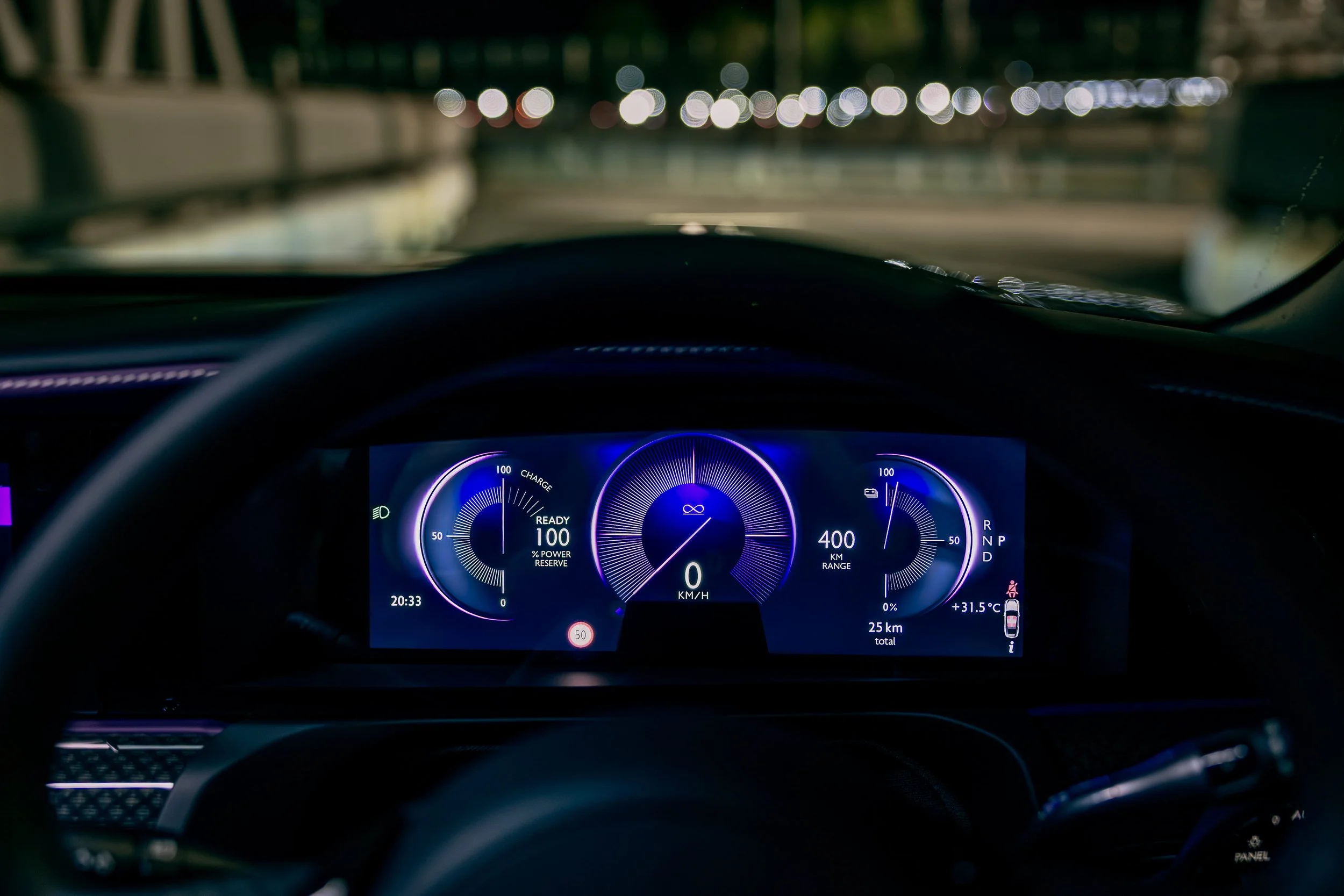 Digital car dashboard displaying fuel level, speed (0 km/h), range (400 km), total kilometers driven (25 km), outside temperature (+31.5°C), time (20:33), and various indicator icons at night with blurred city lights in the background.
