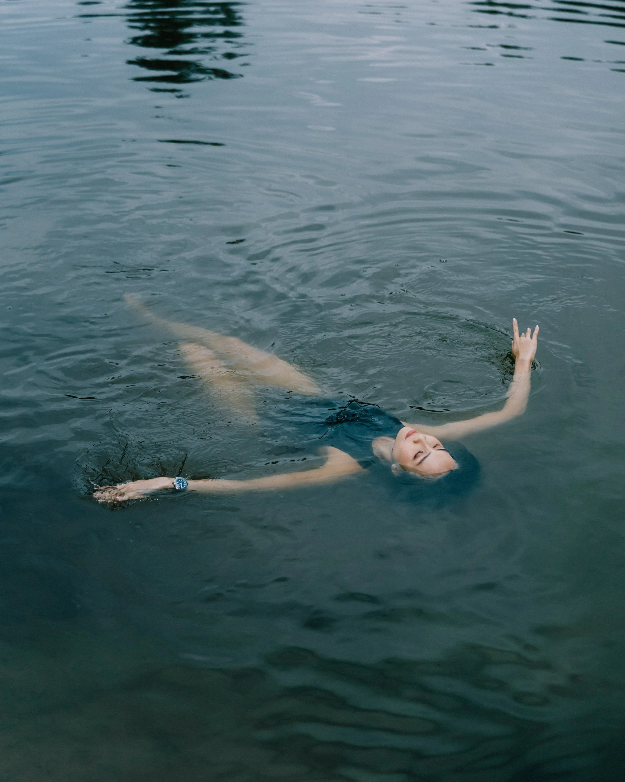 A woman swimming in water, floating on her back with arms extended and eyes closed.