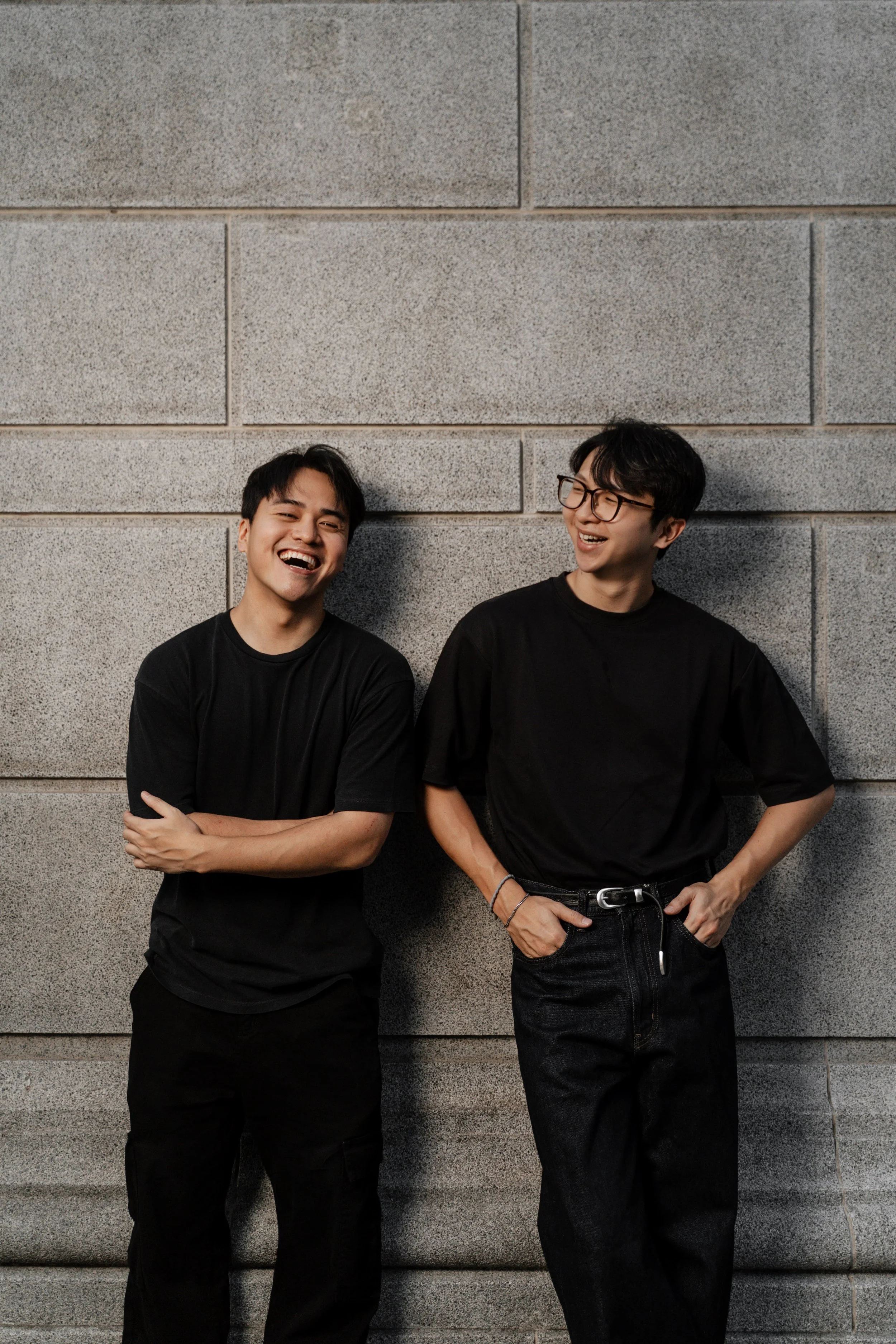 Two young men laughing and leaning against a grey concrete wall, both dressed in black T-shirts and black pants.