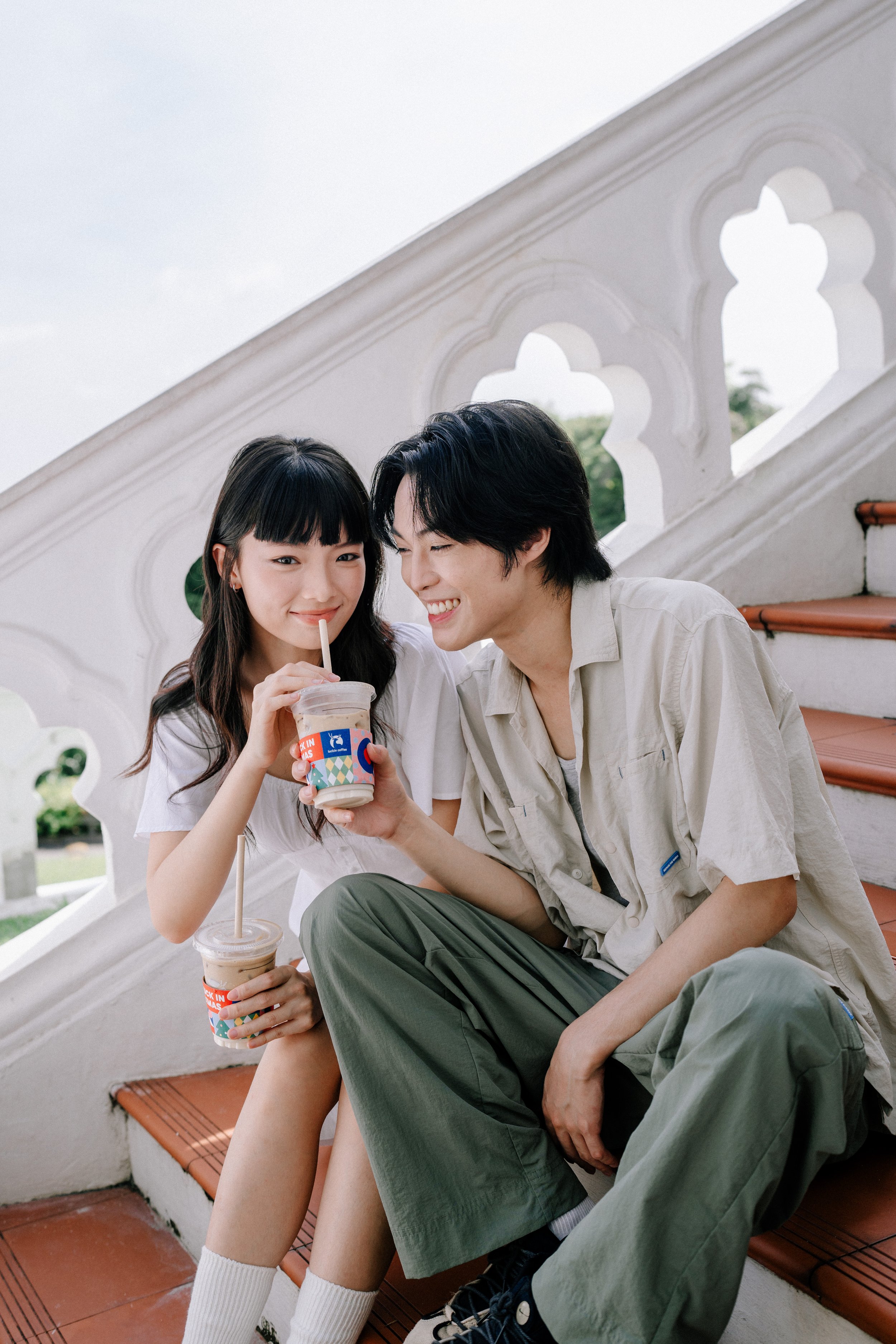 Two young people sitting on outdoor stairs, sharing a moment with one holding a drink, smiling and laughing.