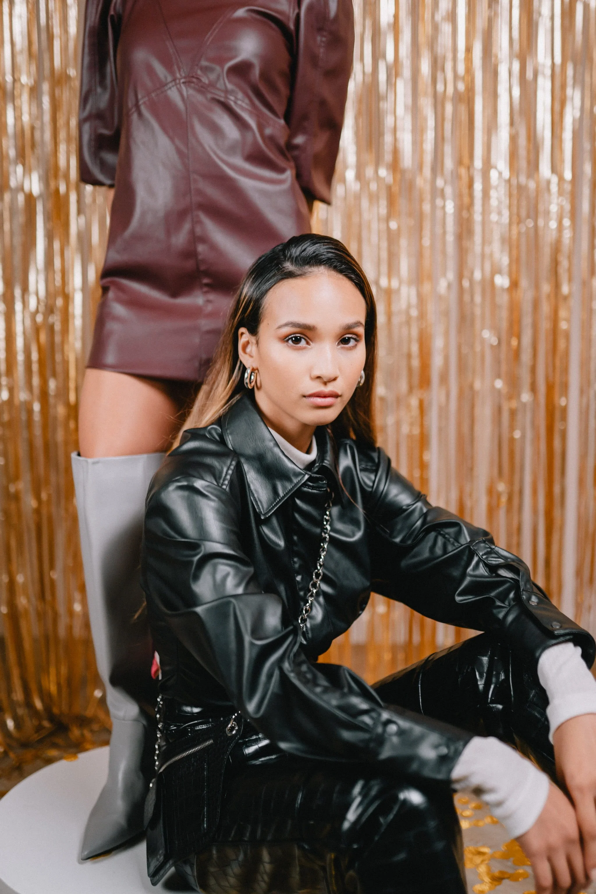 A young woman with long hair wearing a black leather jacket, sitting on a white chair with her arms resting on her knees. A second person, standing behind her, is wearing a maroon leather dress and thigh-high boots. The background features vertical gold-colored curtains.