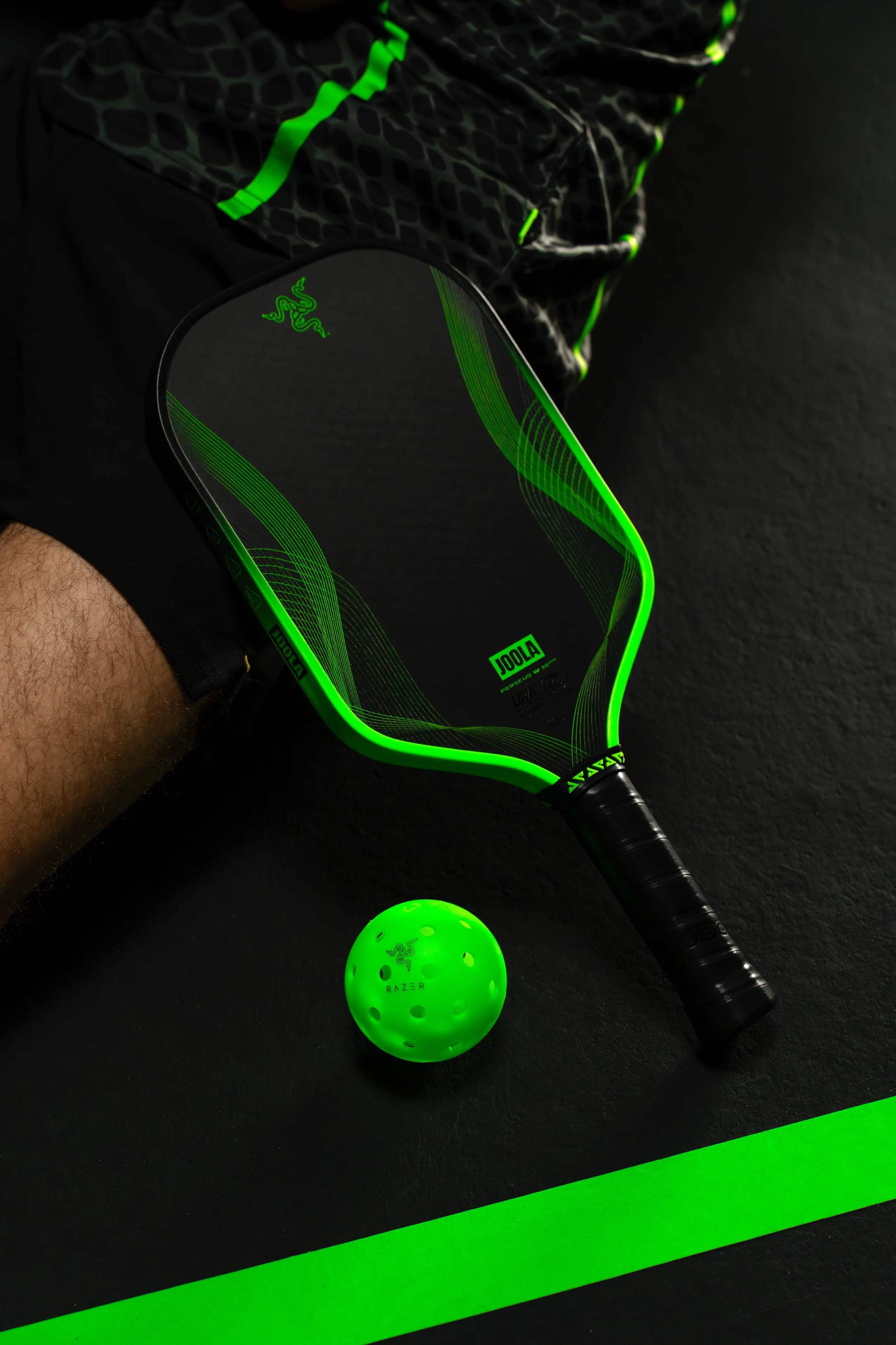 A black and green Razer paddle ball with a small green Razer ball in front, on a black surface with a green line, with a person wearing black and green sportswear in the background.