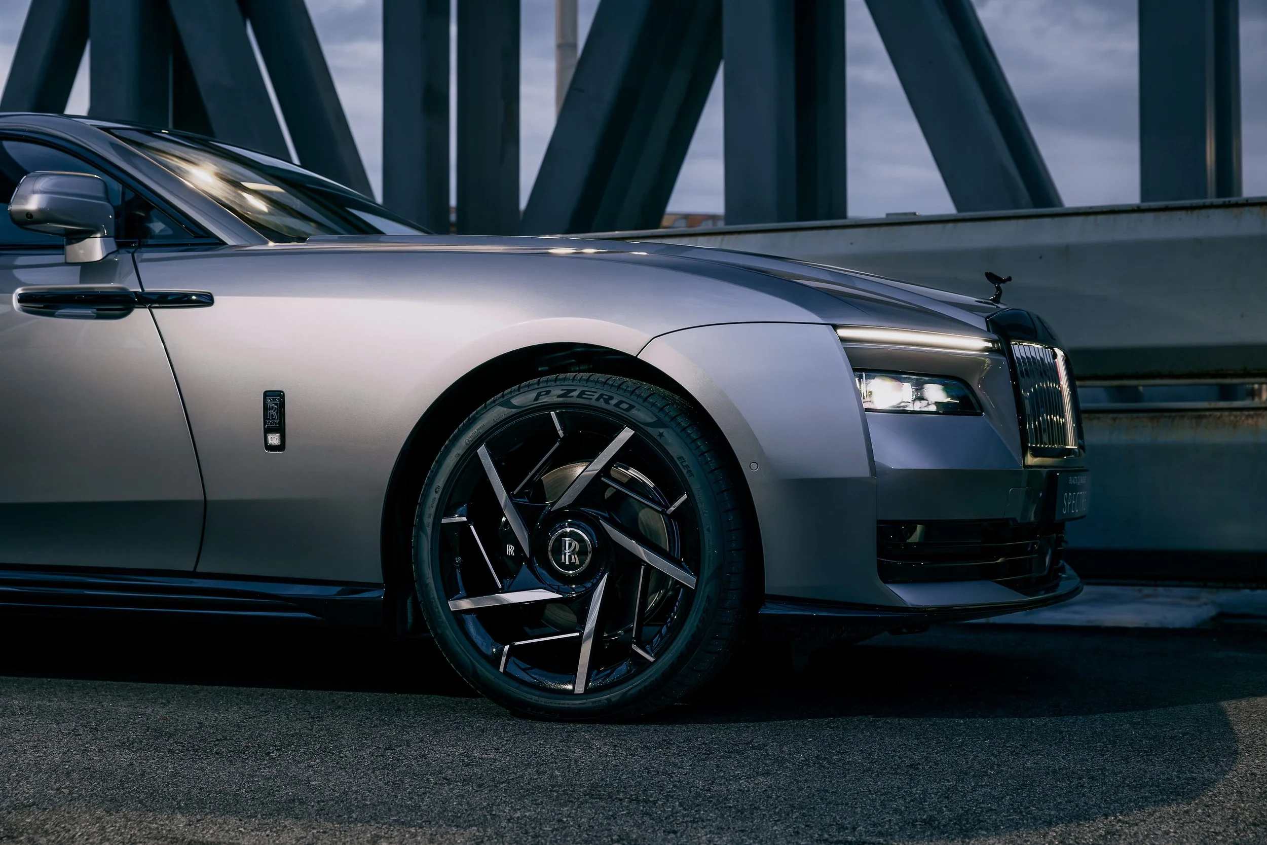 A silver luxury car, possibly a Rolls-Royce, parked on a city street with a modern bridge in the background.