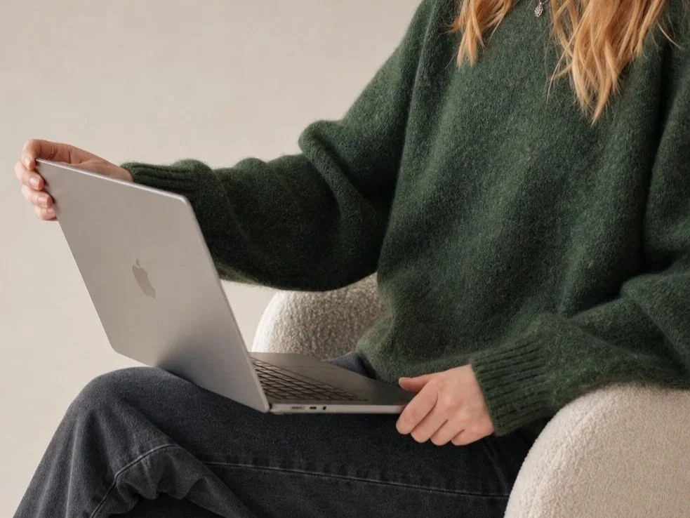 Woman with red hair sitting on a beige chair, holding and using a silver Apple MacBook laptop, wearing a green sweater and dark jeans.