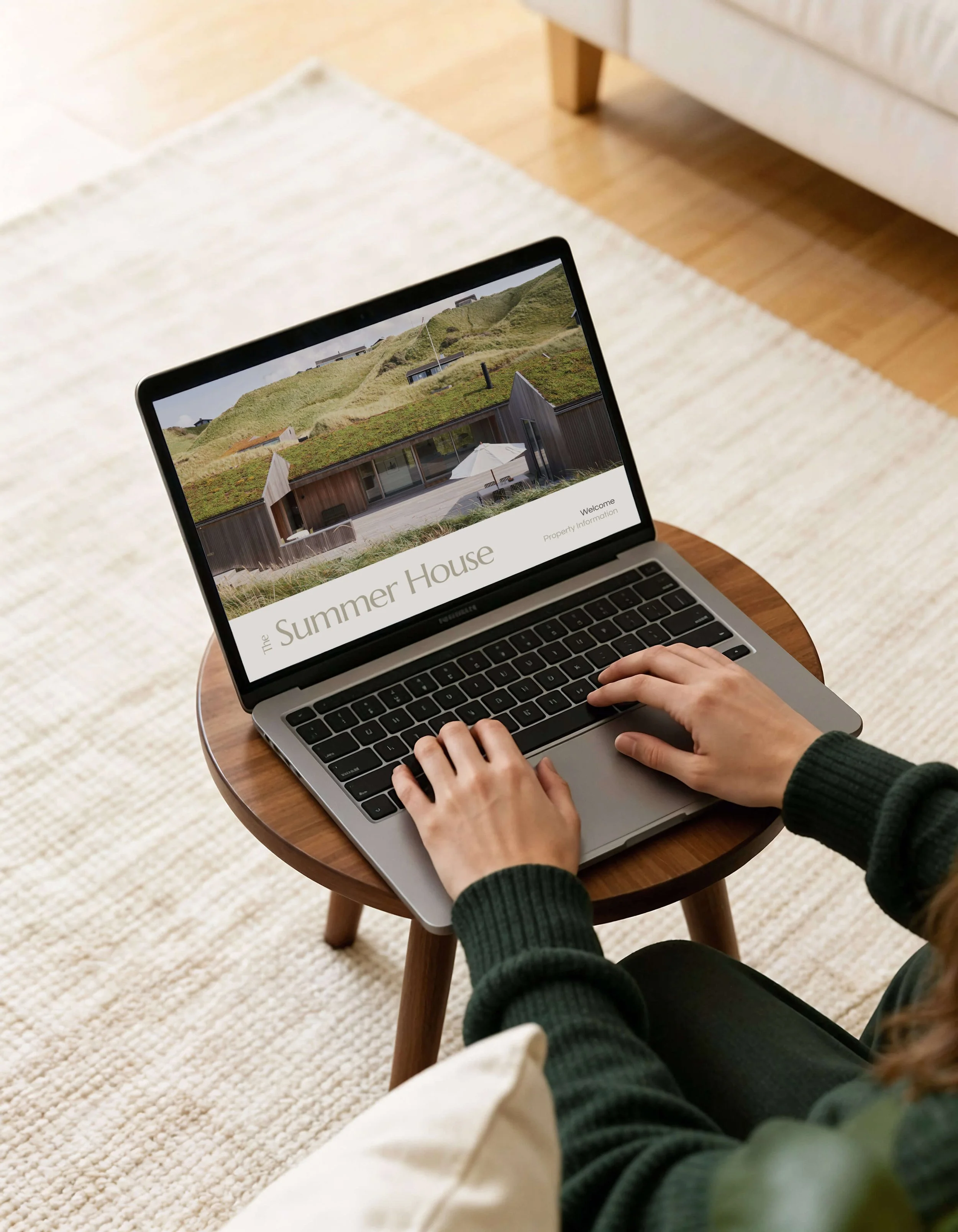 A person sitting at a small round wooden table using a laptop displaying a website about a modern house with green hills in the background.