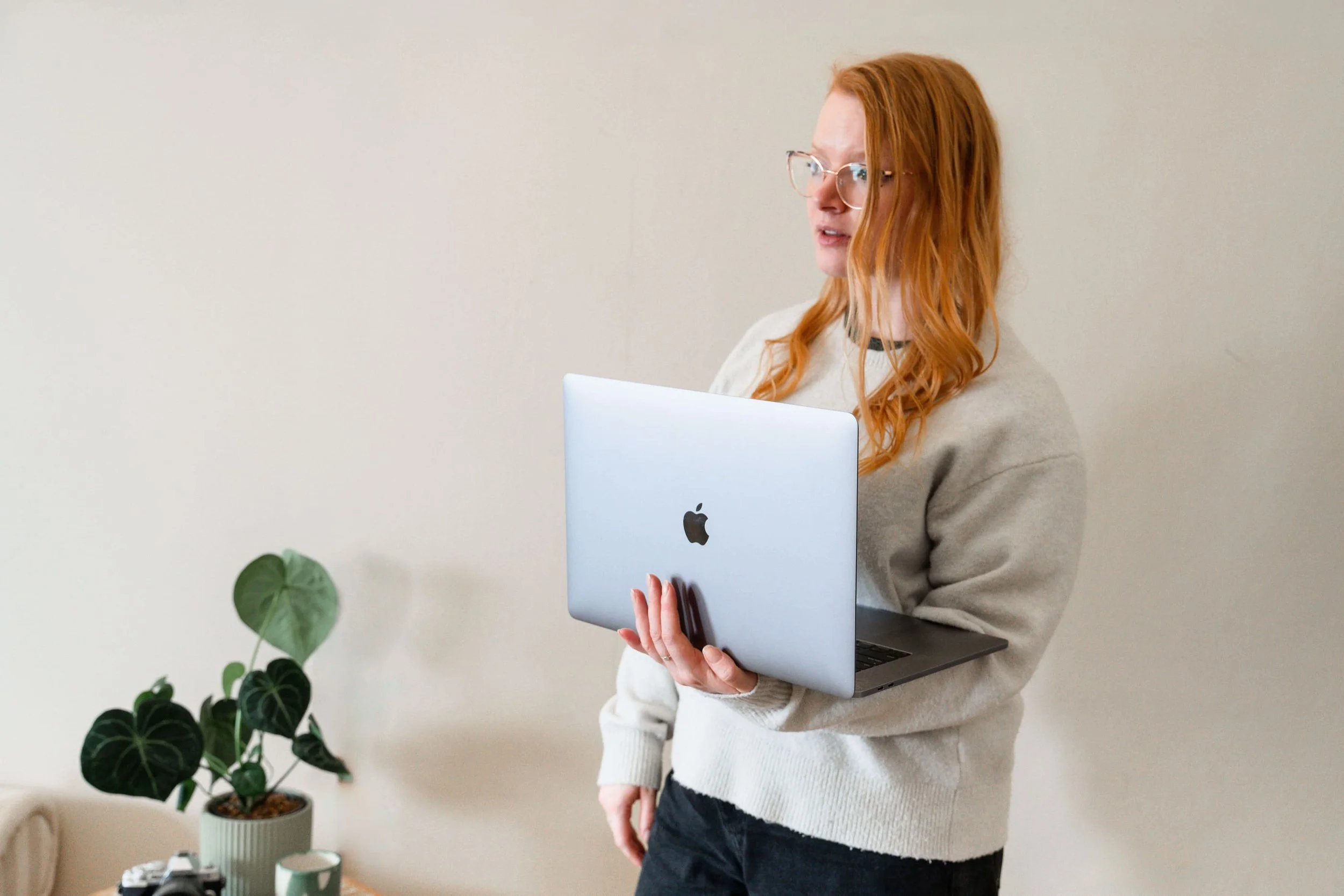 A woman with red hair and glasses holding a silver MacBook laptop in a room with a white wall, a green potted plant, some cameras, and a small round green container on a table.