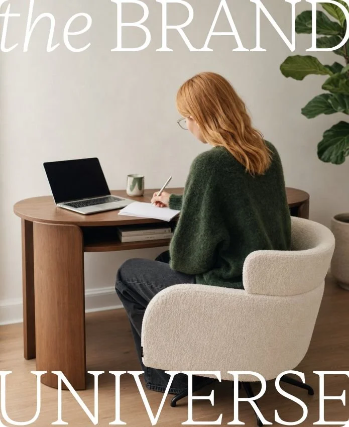 A woman sitting at a wooden desk with an open laptop, writing in a notebook, with a mug nearby, in a room with white walls and a large green plant.