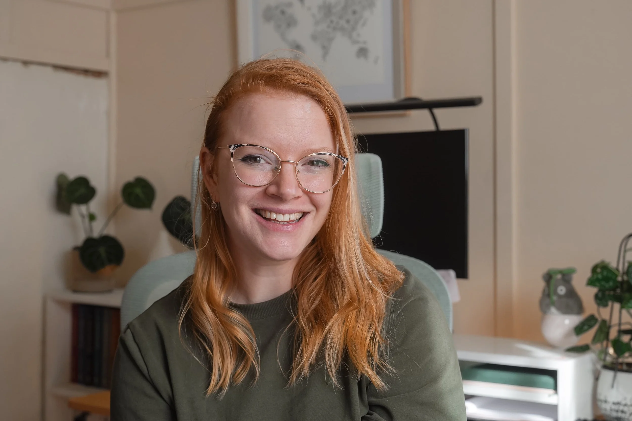 A young woman with red hair, glasses, and earrings smiling in a home or office setting, with plants and a world map in the background.
