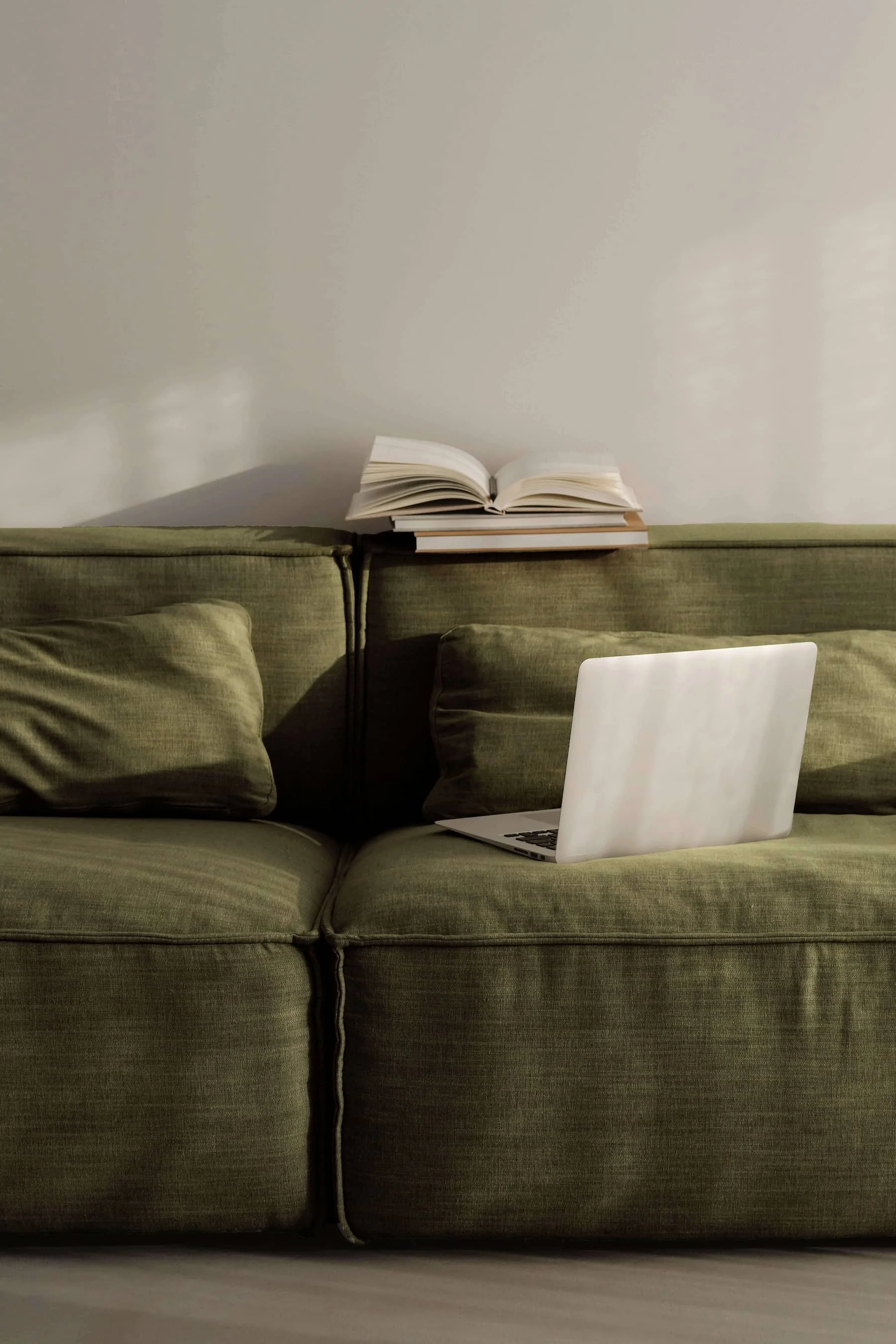A green sofa with pillows, a laptop, and a stack of books topped with an open book, against a plain white wall.