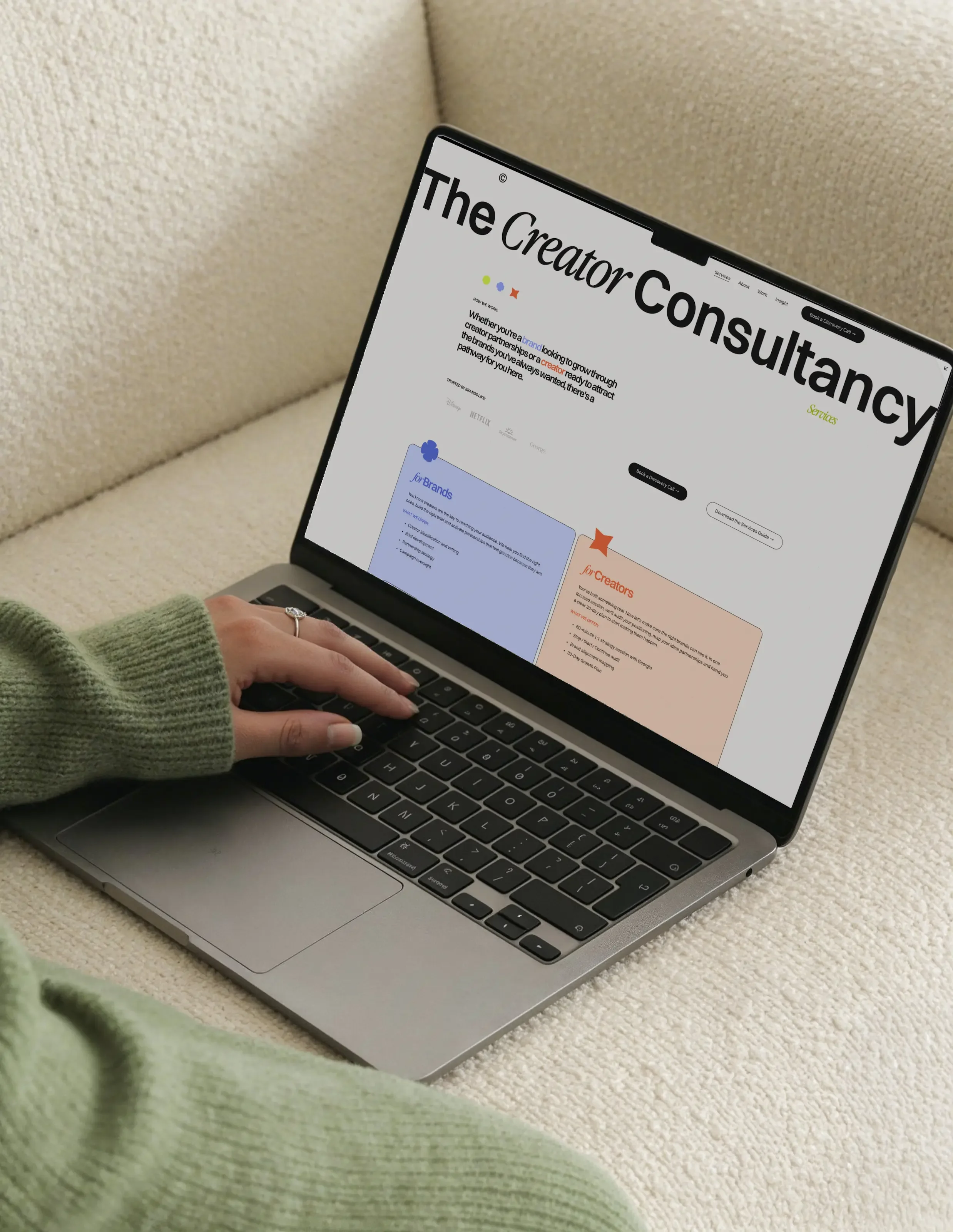 A laptop on a cream-colored surface displaying a website with the heading 'The Creator Consultation.' There is a person's left hand with a ring on the ring finger near the keyboard, and a green fabric item is in the lower left corner.