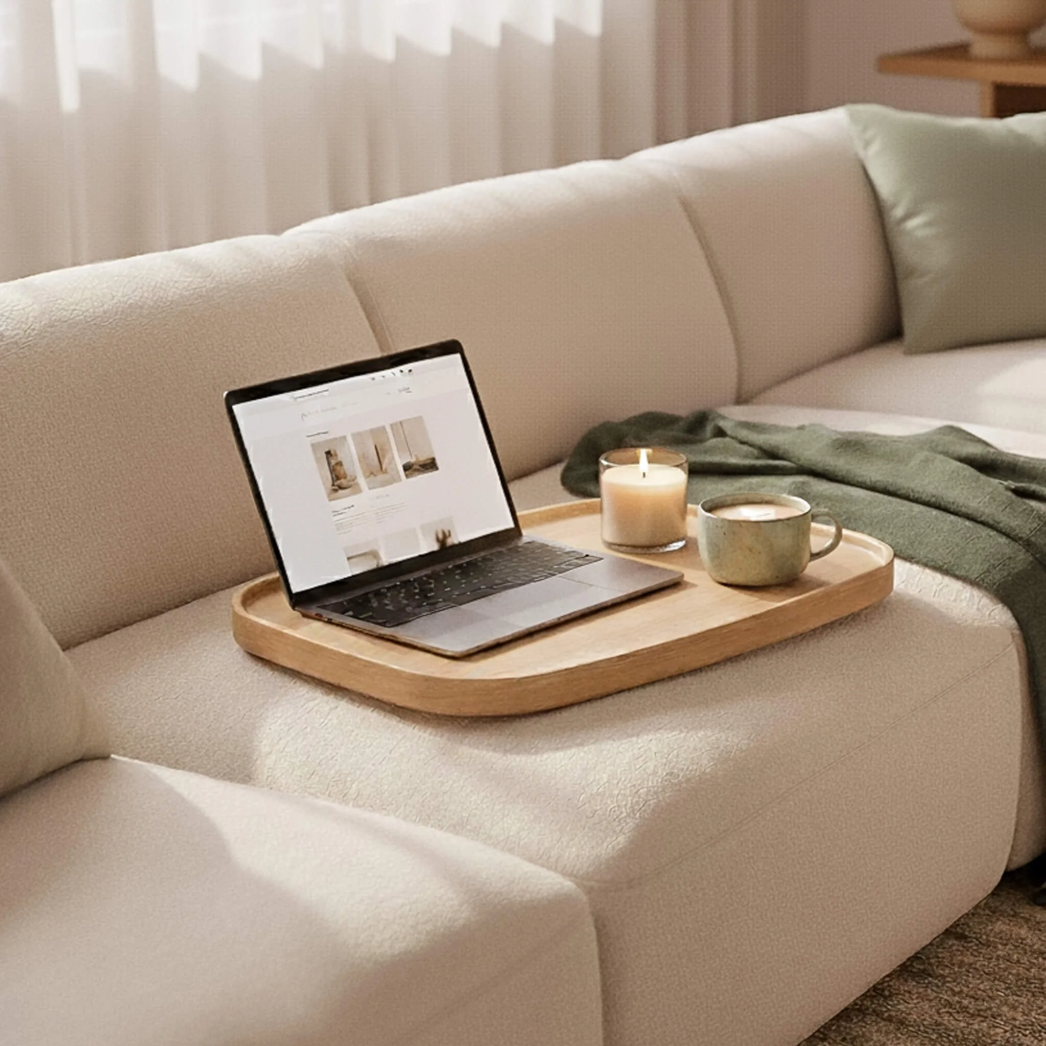 A beige sofa with a tray holding a laptop, a lit candle, and a mug, next to a green blanket and pillows in a cozy living room.