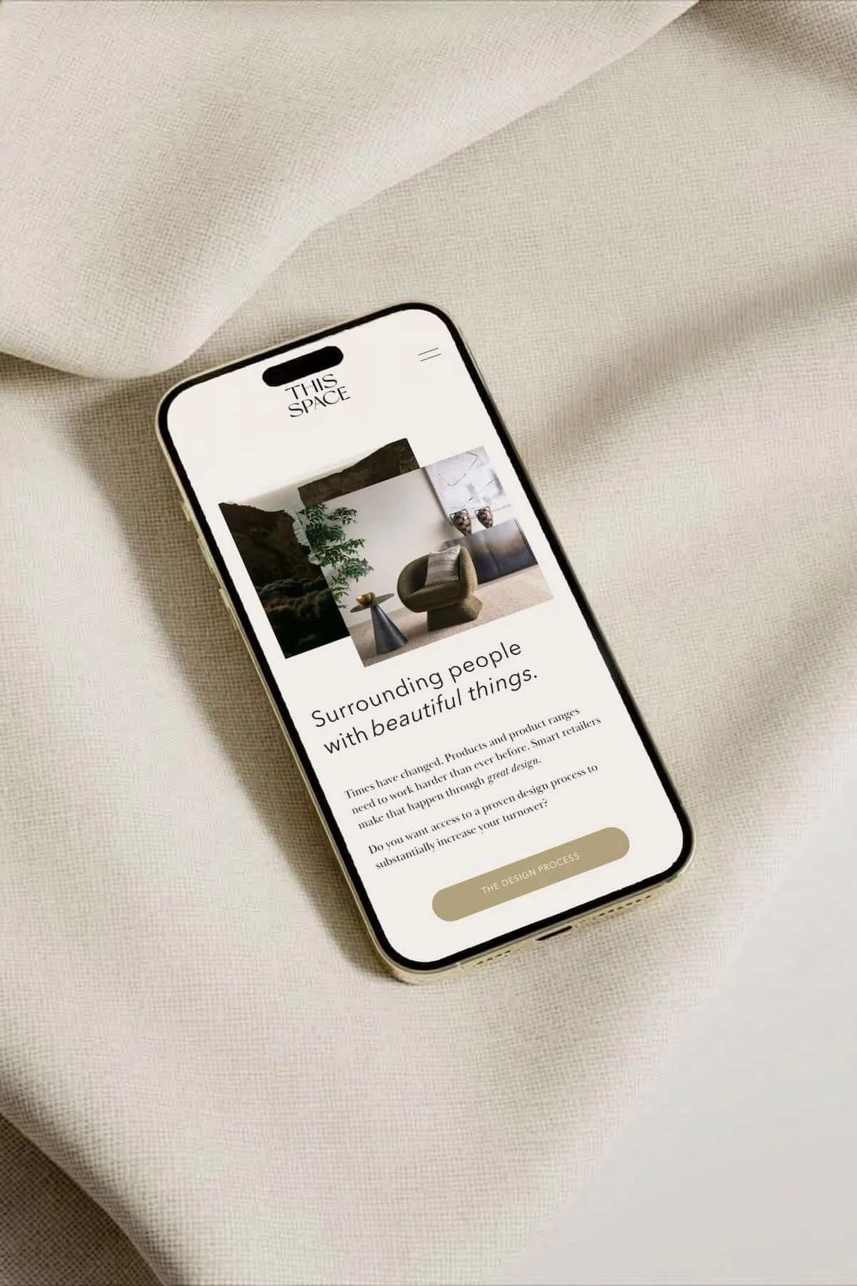 Smartphone displaying a website titled "This Space," with an image of modern living room furniture and a section titled "Surrounding people with beautiful things." The phone is placed on a light-colored, textured fabric surface.