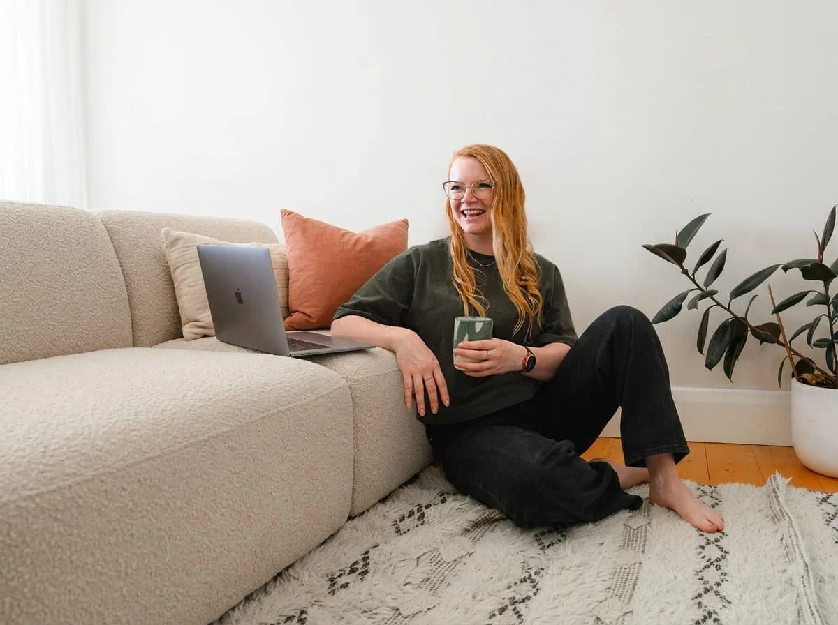 A woman with red hair and glasses sitting on the floor next to a beige sofa, holding a mug, with a laptop and pillows nearby, a potted plant in the background.