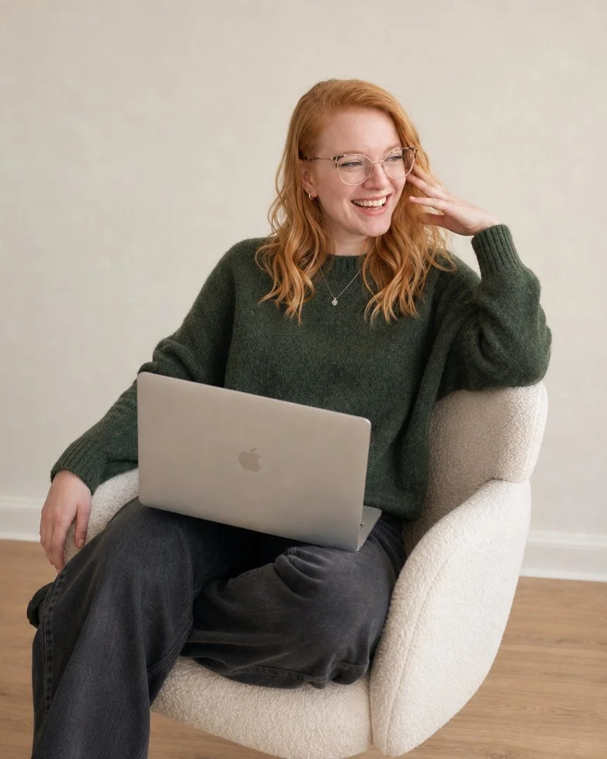 A young woman with red hair, glasses, and a dark green sweater sitting on a white armchair with a laptop on her lap, smiling and laughing with one hand touching her face.
