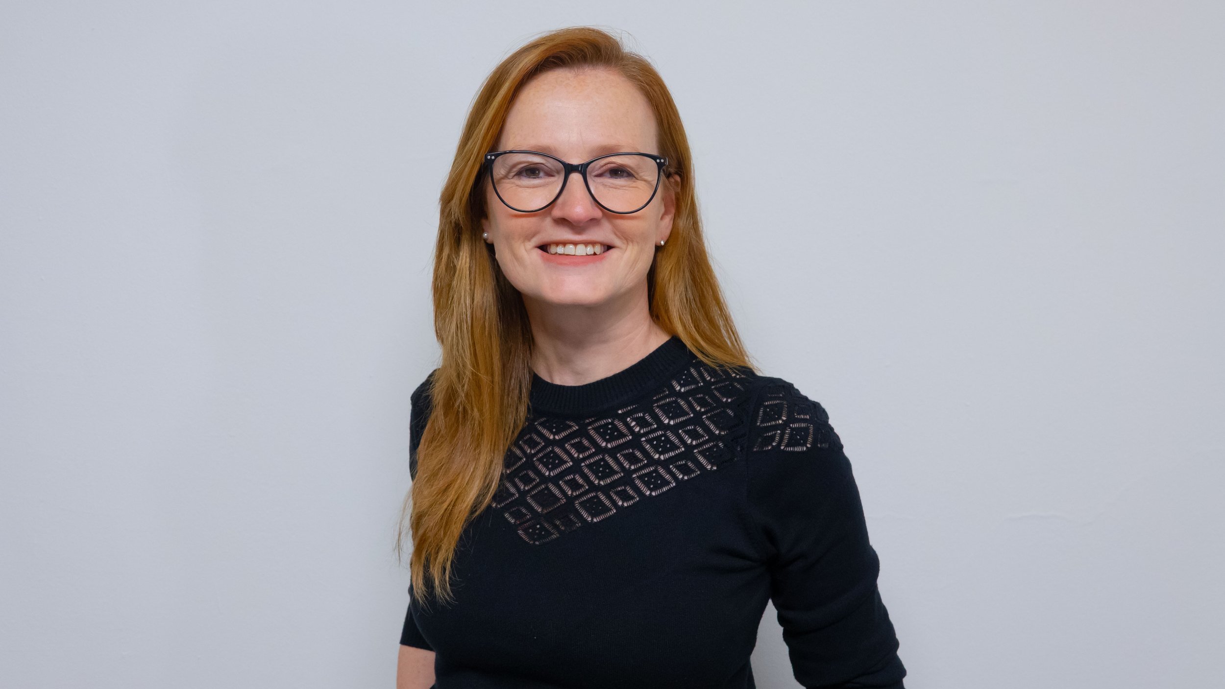 Mujer con cabello largo y rojo, gafas negras, camiseta negra con diseño geométrico, sonriendo, fondo gris.