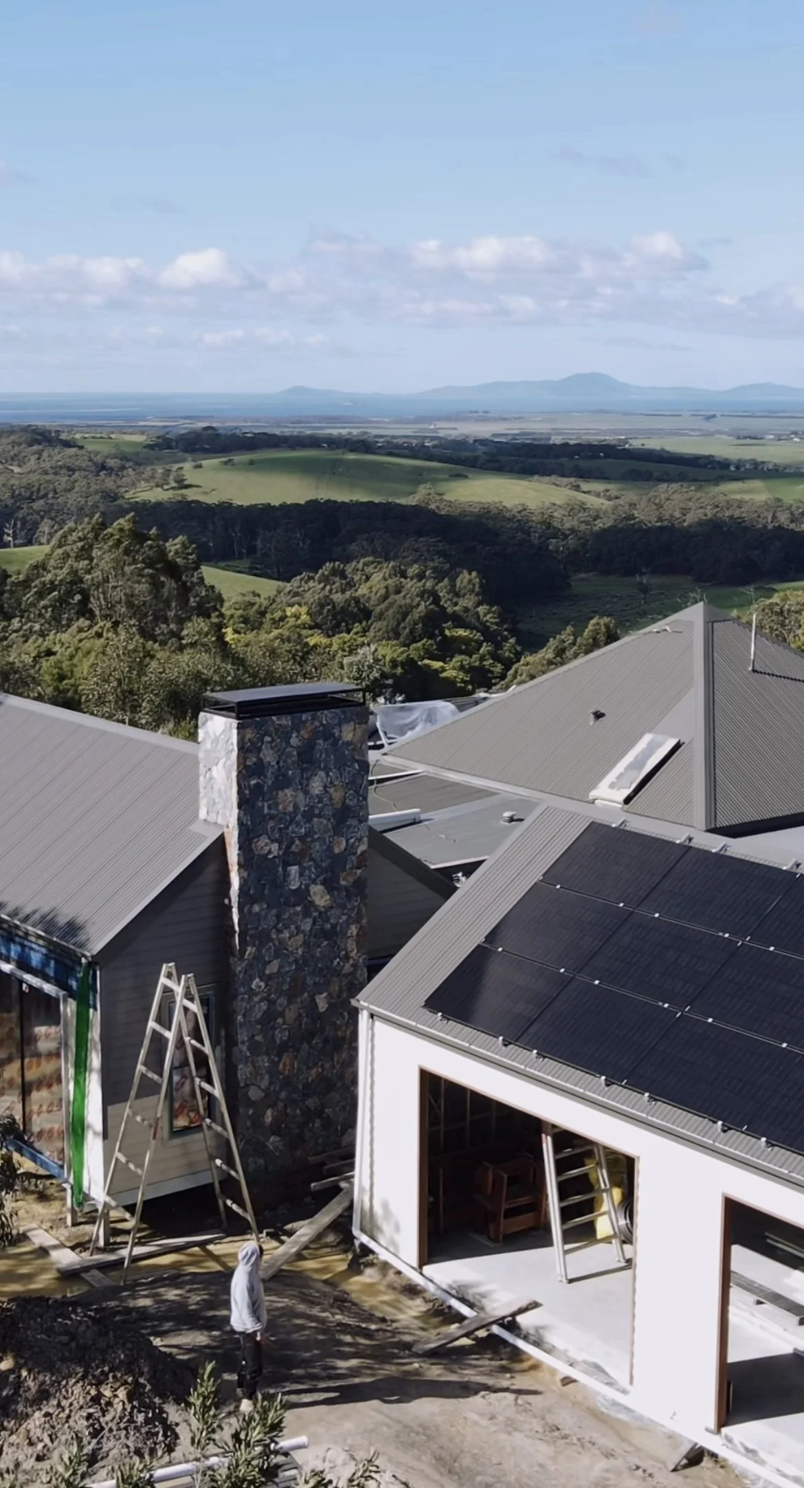Construction site with partially built houses, ladders, and a person in a white hoodie, overlooking a green landscape with hills and blue sky.