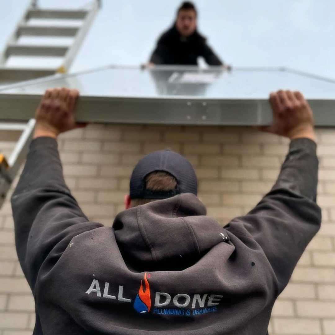 A worker in a black hoodie with the logo 'All Done Plumbing & Drainage' is lifting a large metal panel or roofing sheet above his head. Another person, standing on a ladder, is on a roof in the background, visible from a low angle.