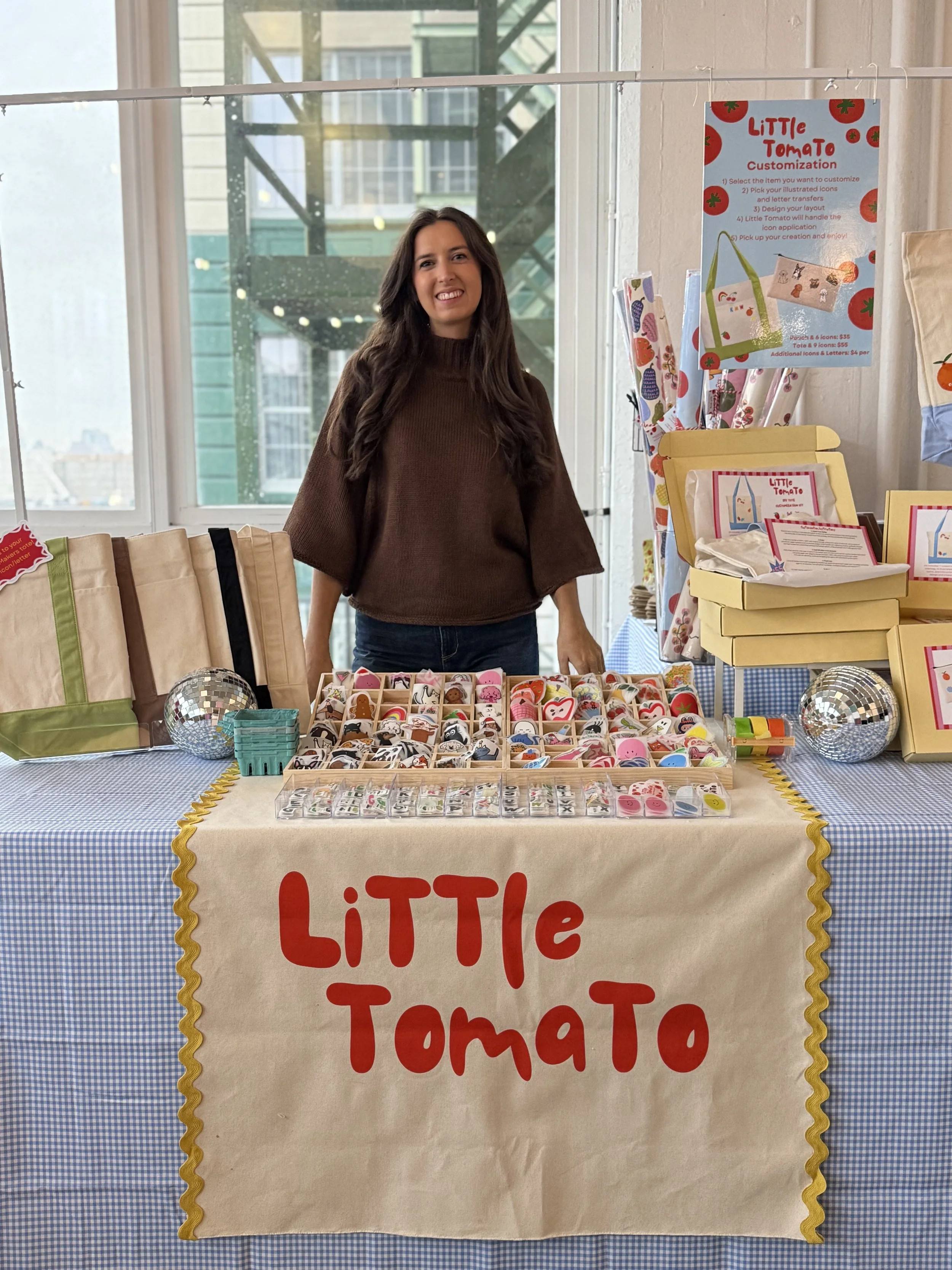 A woman stands behind a booth with a sign that reads 'LITTLE TOMATO.' The booth displays various colorful felt or fabric items, likely and accessories, on the table and in open boxes. The background includes a large window with a cityscape view.