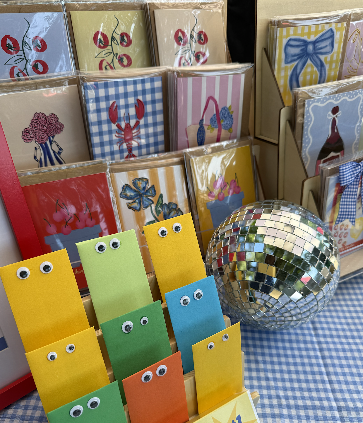 Colorful greeting cards with illustrations of cherries, lobsters, flowers, and bows displayed in a store. Small paper notebooks with googly eyes resembling eyes are in the foreground, and a disco ball is on the table with a blue and white checkered t