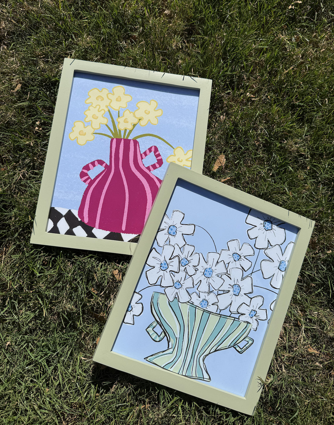 Two framed illustrations of flower bouquets placed on grass. One shows a pink striped vase with yellow flowers against a blue background, the other depicts a striped green and blue vase with white flowers featuring blue centers against a blue backgro
