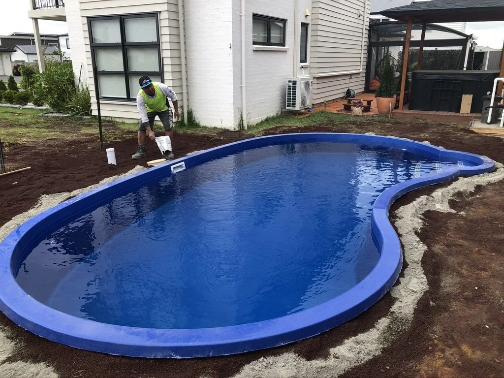 A person in a neon vest and hat is working near a newly installed kidney-shaped blue swimming pool in a backyard, with some gardening tools and dirt around the area.