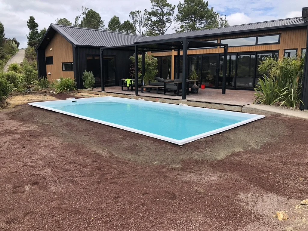 A backyard with a rectangular in-ground swimming pool, a black and wood house with a covered patio, and surrounding greenery.