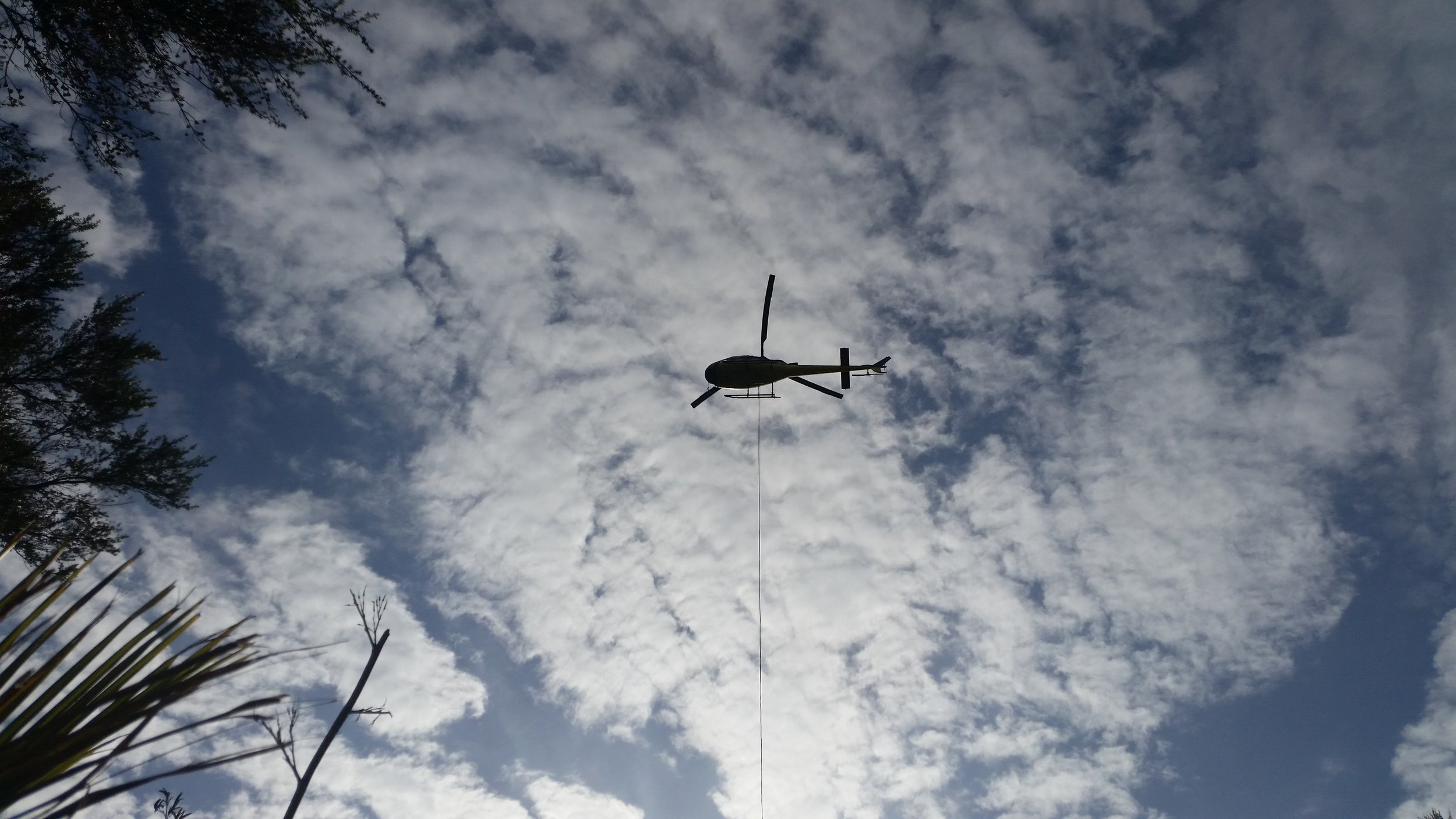 A helicopter flying in the sky with scattered clouds, trees visible at the edges of the image.